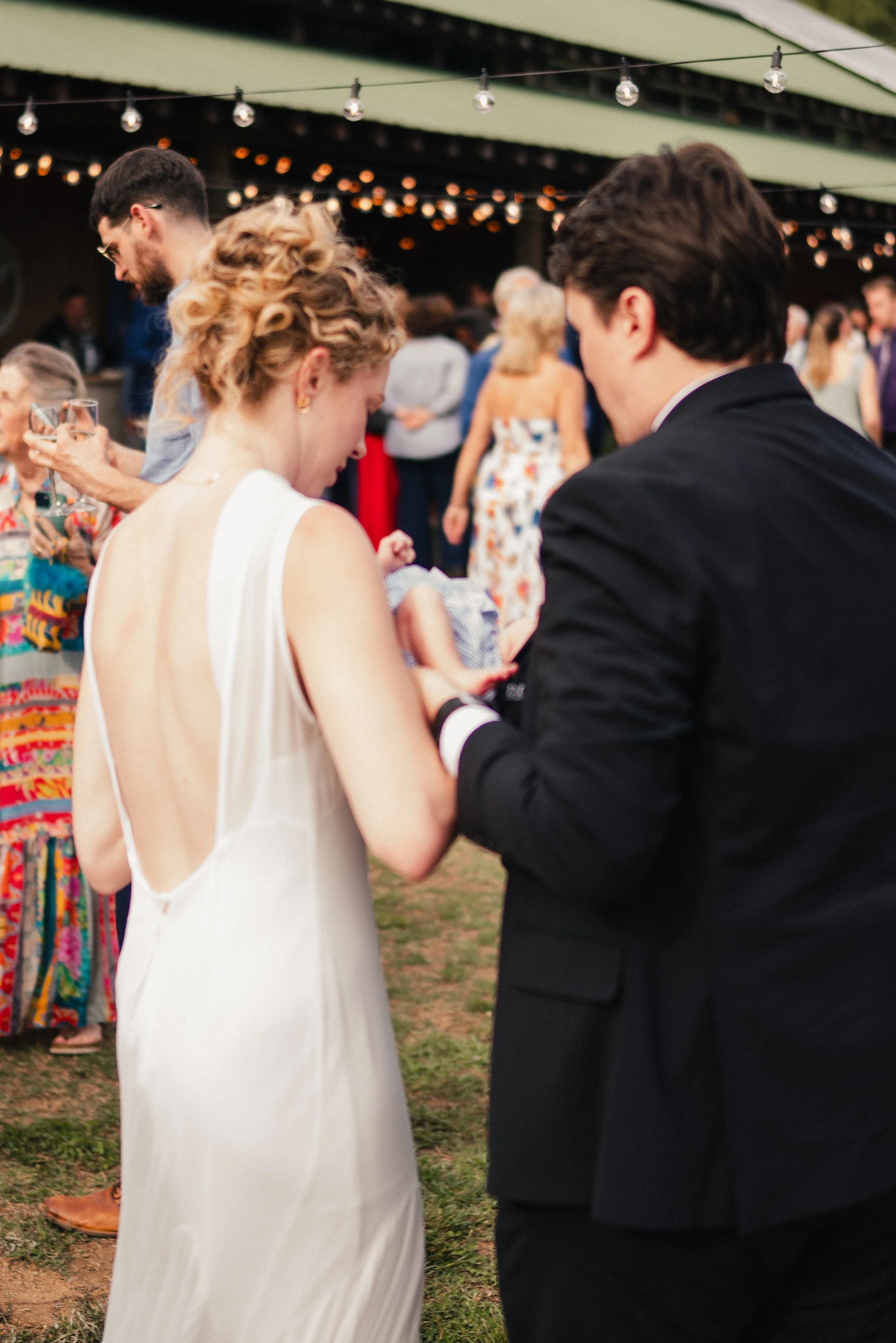A bride and groom at a wedding ceremony, holding hands and looking at each other, surrounded by guests under string lights and outdoor decorations.