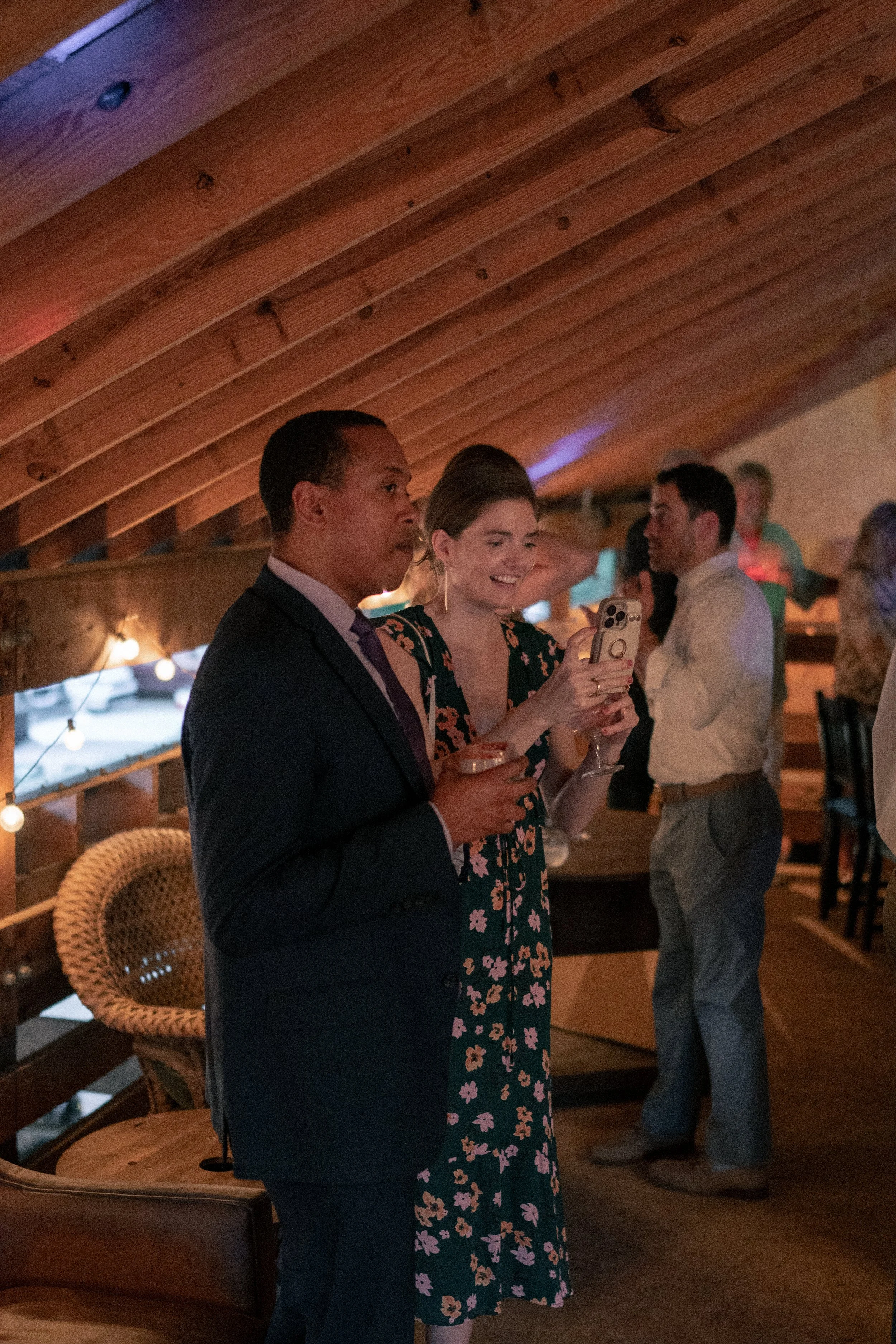 A group of people at a social gathering inside a wooden cabin with a curved wooden ceiling. Two people are prominently in the foreground, one man in a suit and a woman in a floral dress, both holding drinks and smiling. Other individuals are in the b