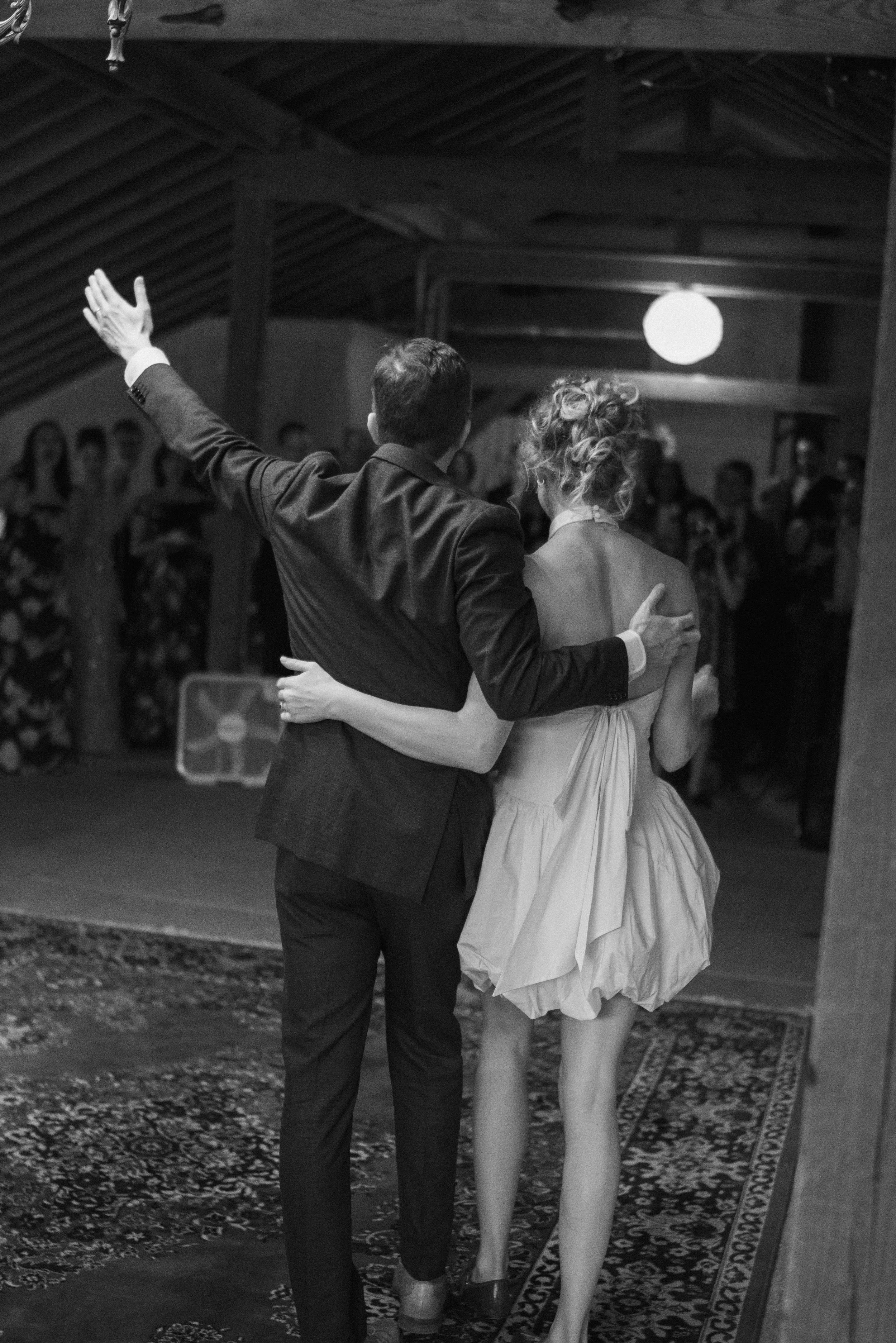 A couple, a man in a suit and a woman in a dress, dance closely together in an indoor setting with a patterned rug on the floor. The man has his arm around the woman, and she is holding his shoulder. The woman's dress is short, and they are facing aw