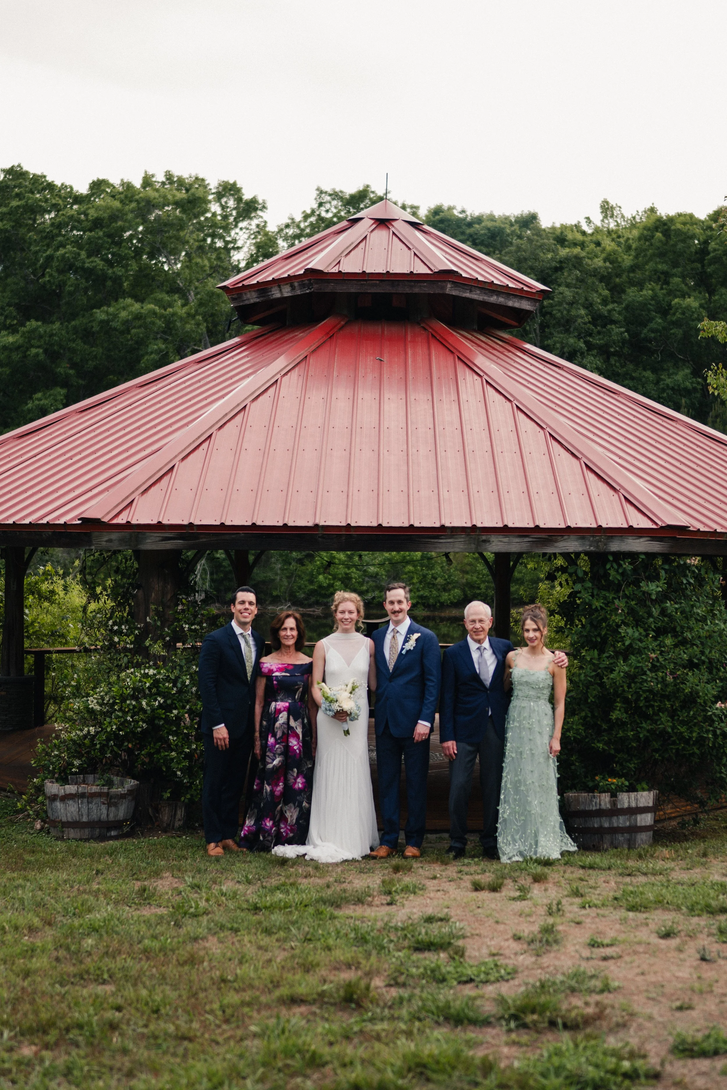 Group of seven people standing in front of a pavilion with a red metal roof, surrounded by trees and greenery, during daytime.