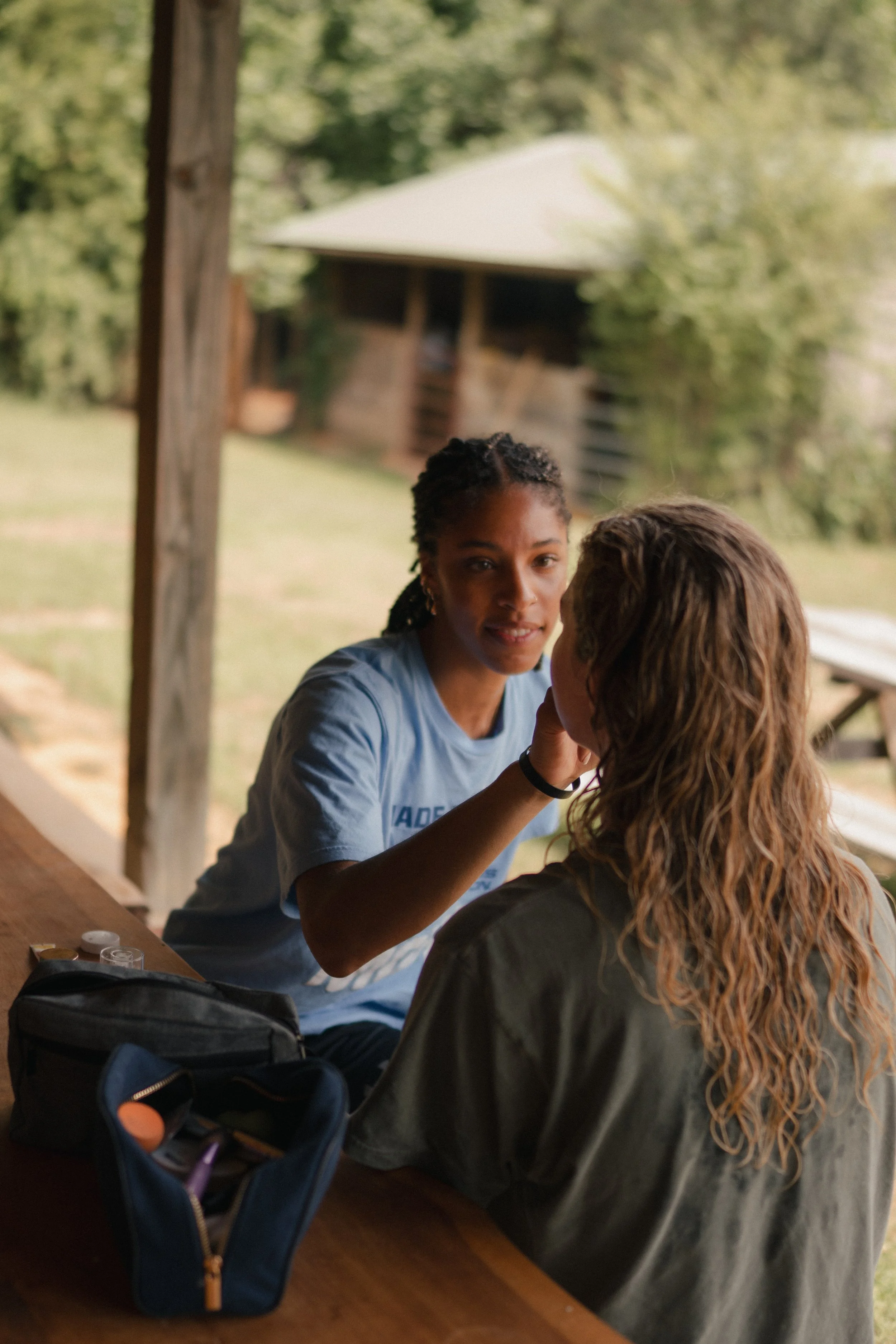 Two young women sitting at a wooden table outdoors, one applying makeup or face paint to the other's face, with a barn and trees in the background.