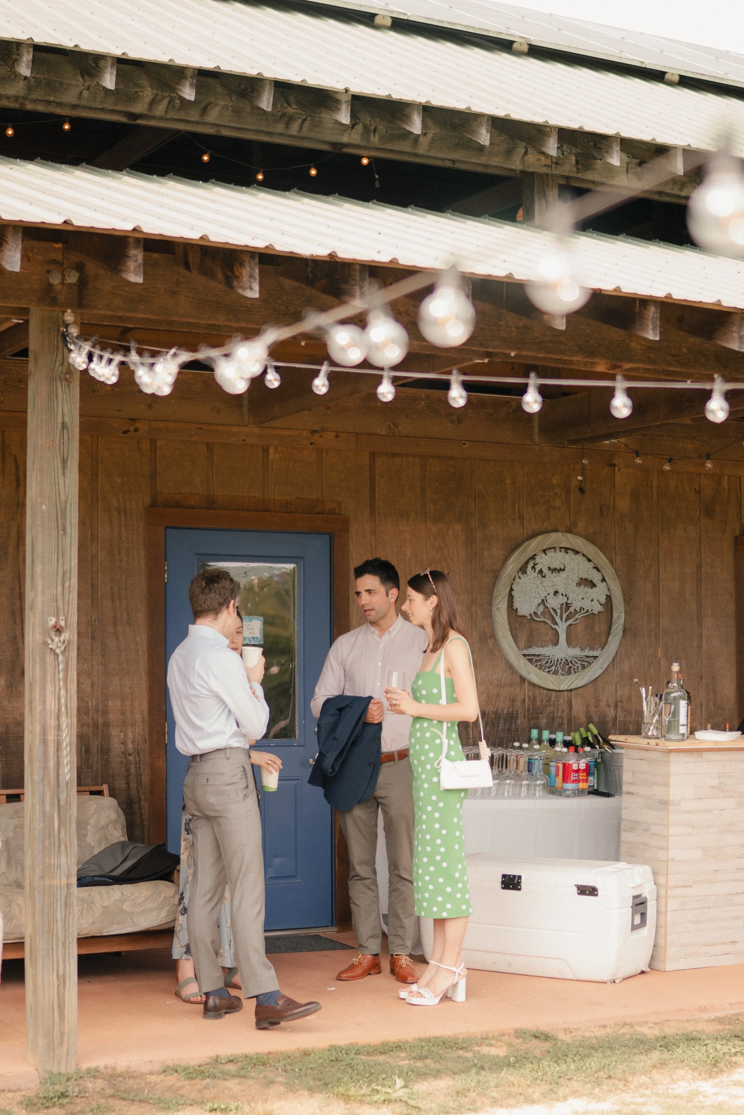 A group of five people socializing at an outdoor event under a wooden structure decorated with string lights. They are dressed in semi-formal attire, with some holding drinks, during daytime.