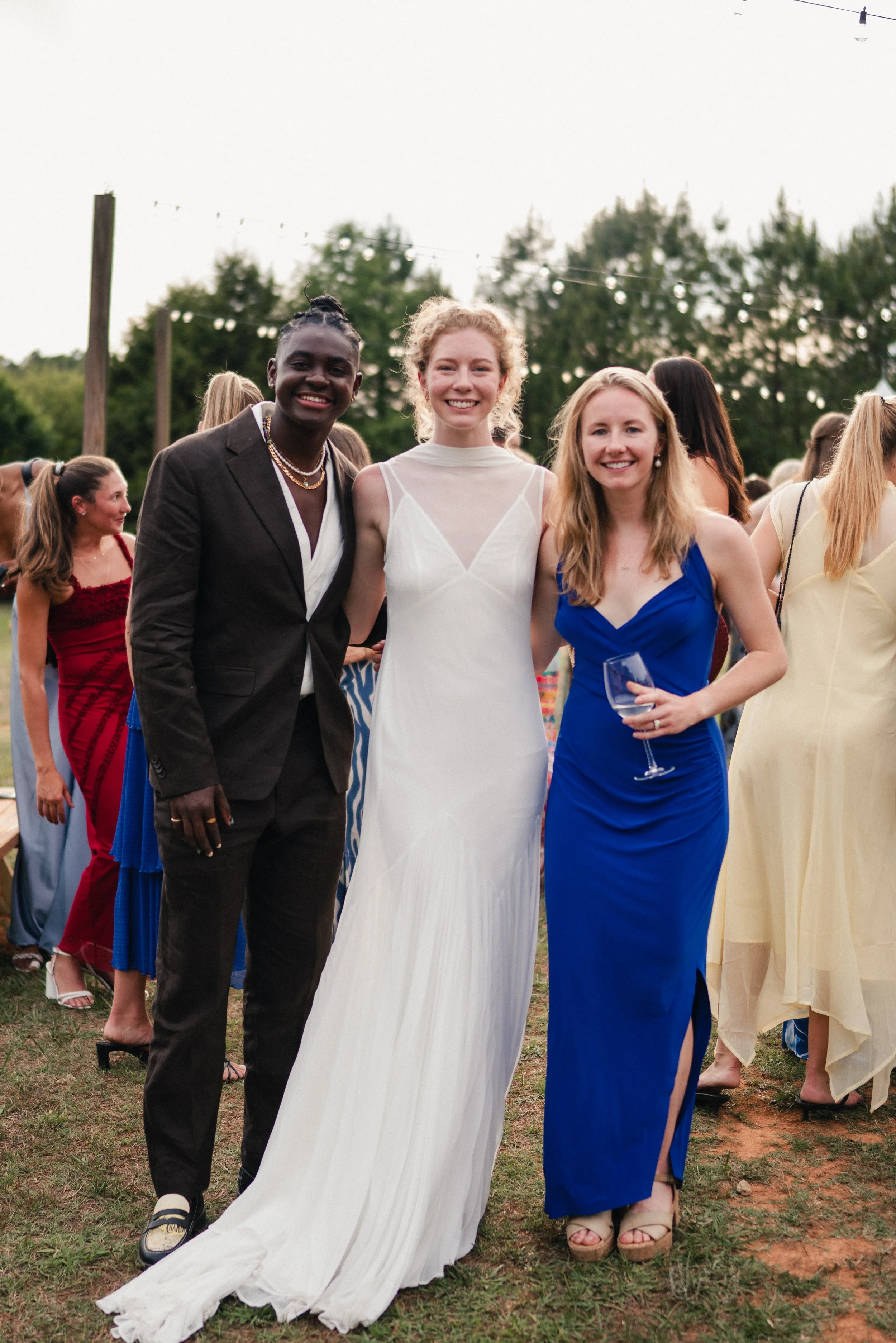 Three women are standing outdoors at a social event, smiling at the camera. One woman wears a white dress, another a blue dress, and the third a black suit with jewelry. People are mingling in the background, and string lights hang overhead during th