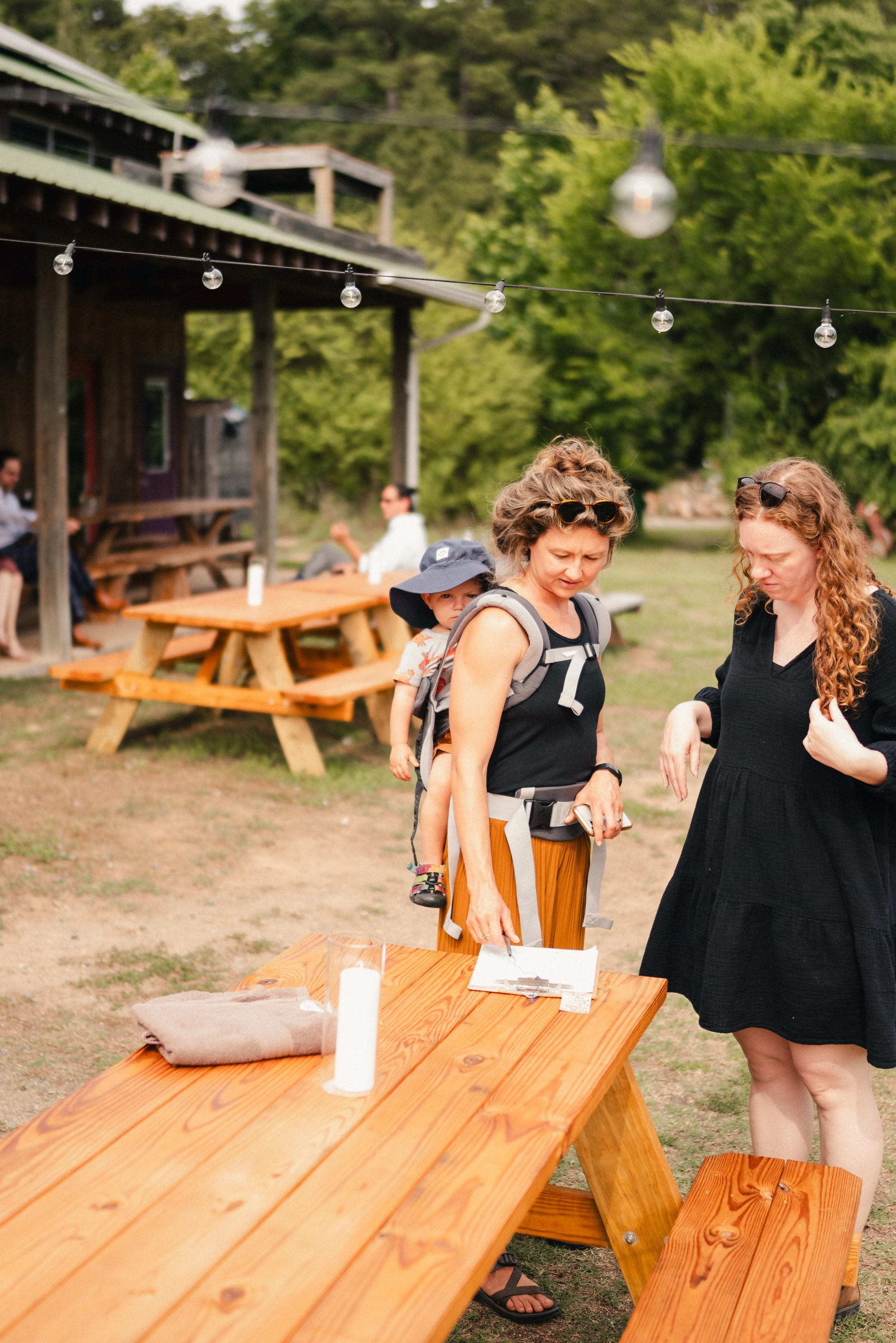 People gathered outdoors near wooden picnic tables, some sitting, some standing, with string lights hanging overhead and surrounded by trees.