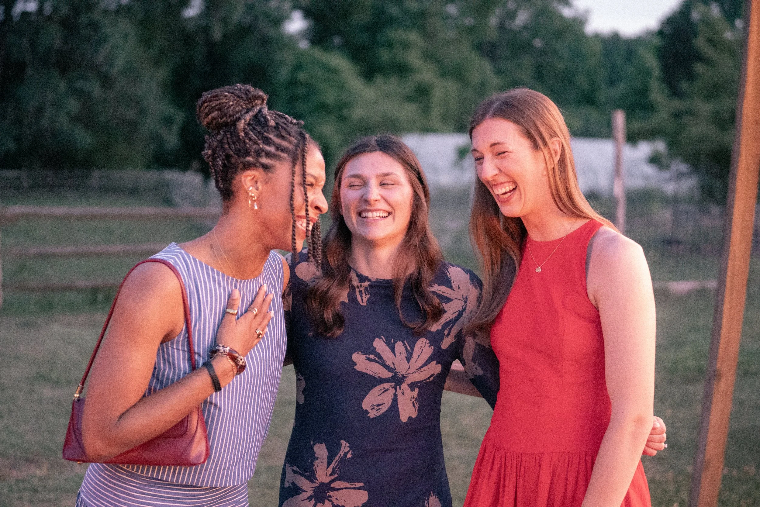 Three women smiling and laughing outdoors, wearing casual summer dresses, standing close together during a warm evening.