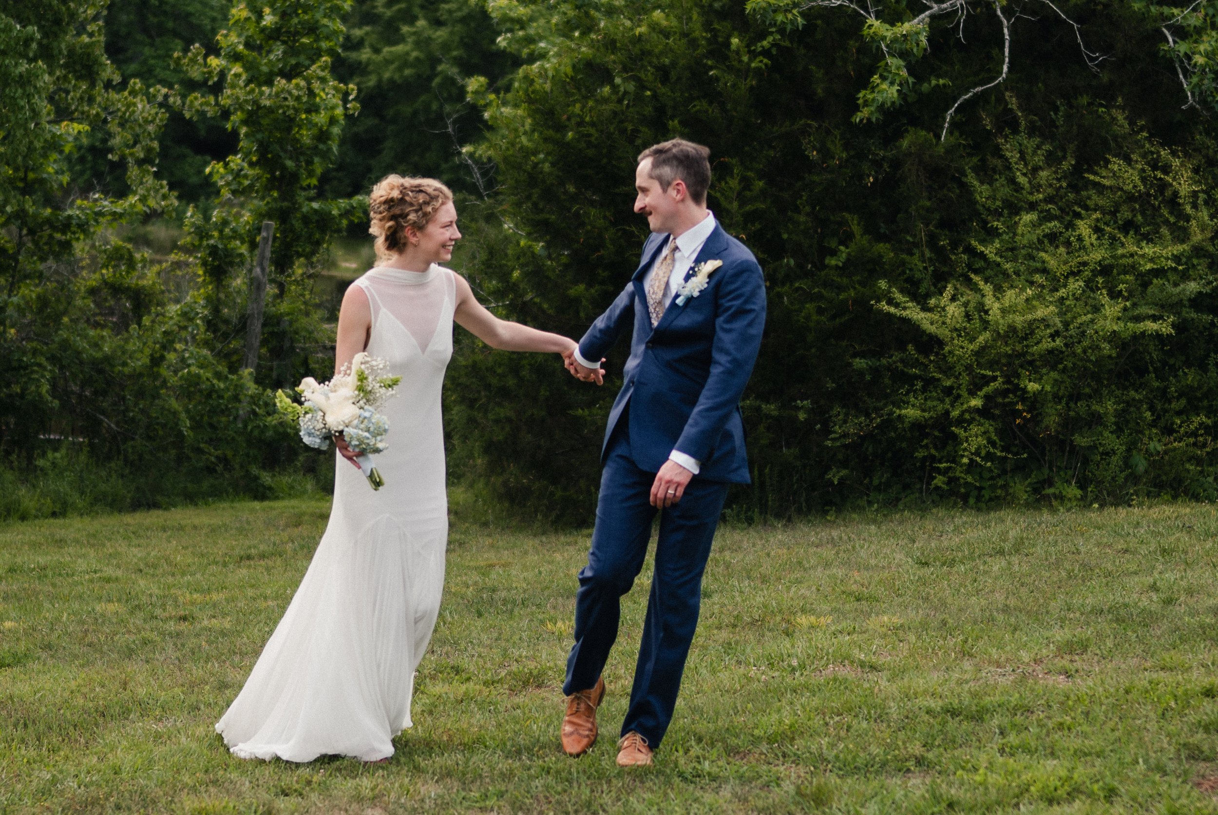 A bride and groom holding hands and smiling outdoors on a grassy field with green trees in the background.