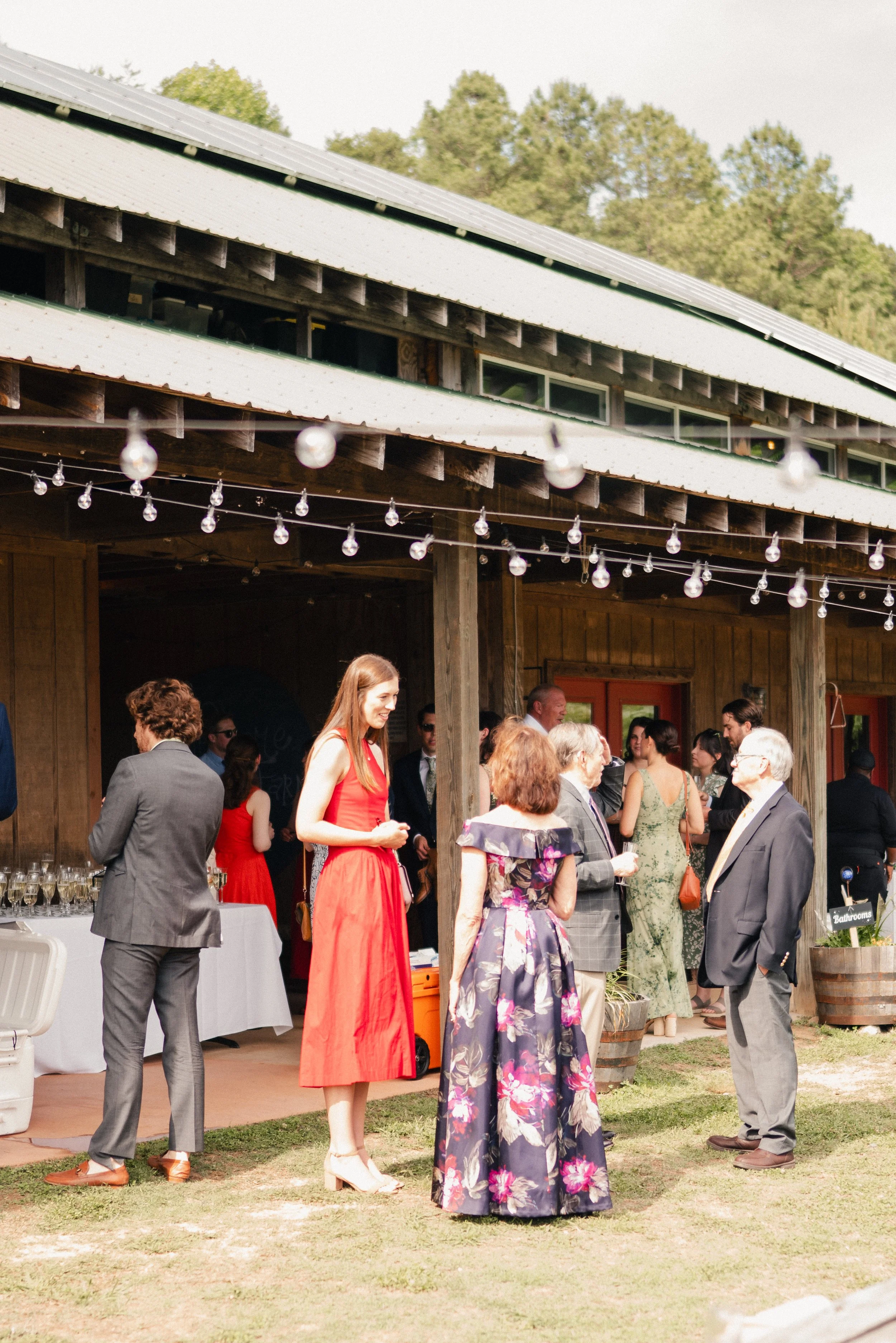 People socializing outdoors at a gathering near a wooden building with string lights, some holding drinks, with trees in the background.