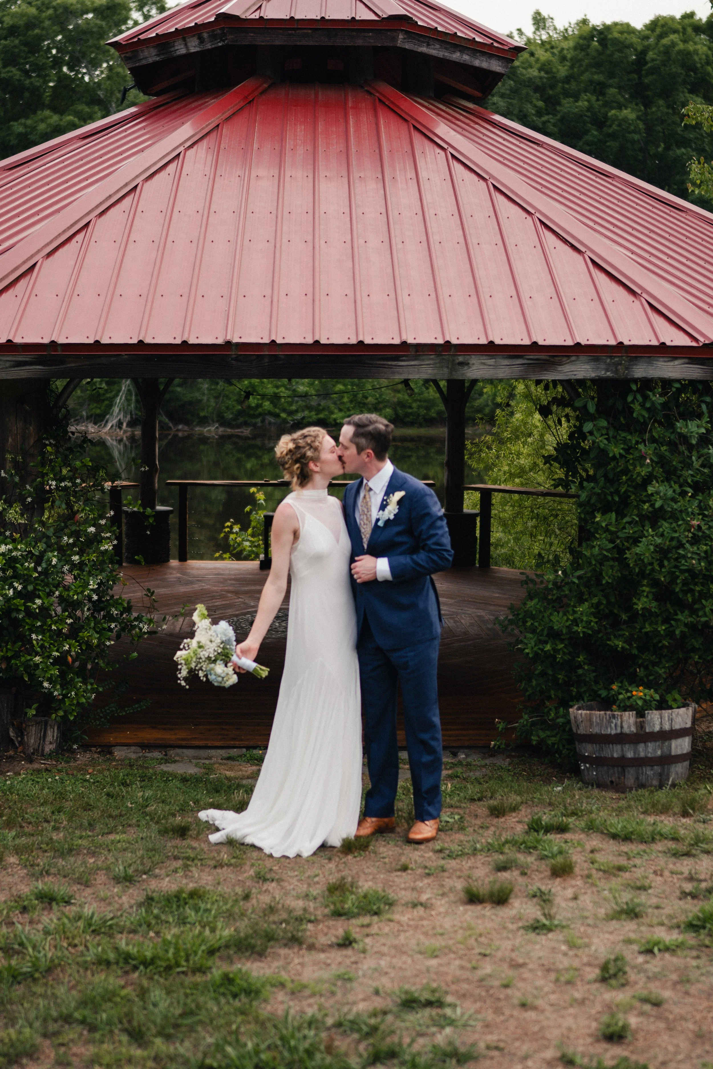 A newlywed couple sharing a kiss outdoors under a red metal roof gazebo, with the bride holding a bouquet and the groom in a blue suit, surrounded by greenery and a rustic setting.