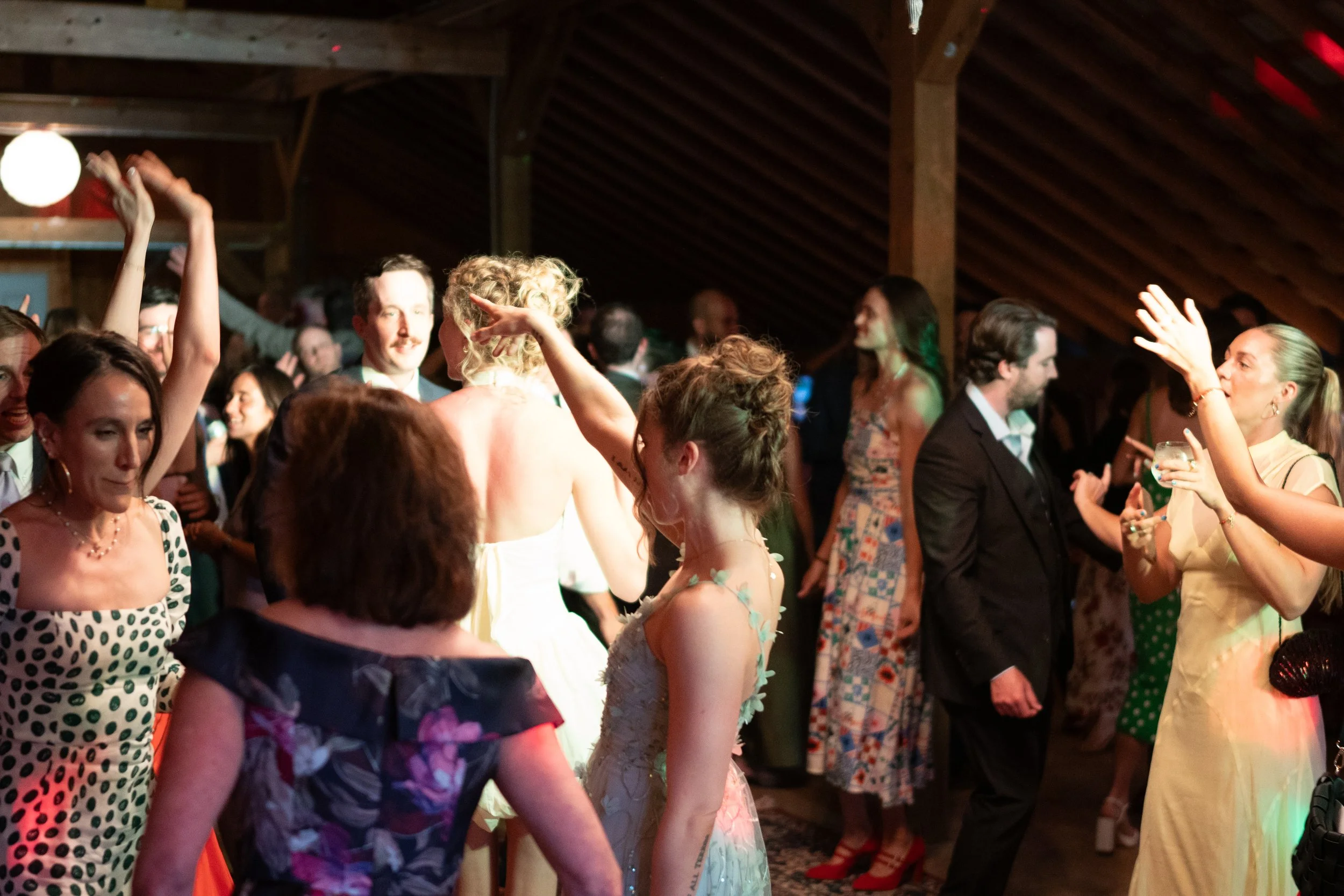 People dancing and socializing at a lively party or celebration in a wooden venue, with some individuals wearing colorful dresses and formal attire.