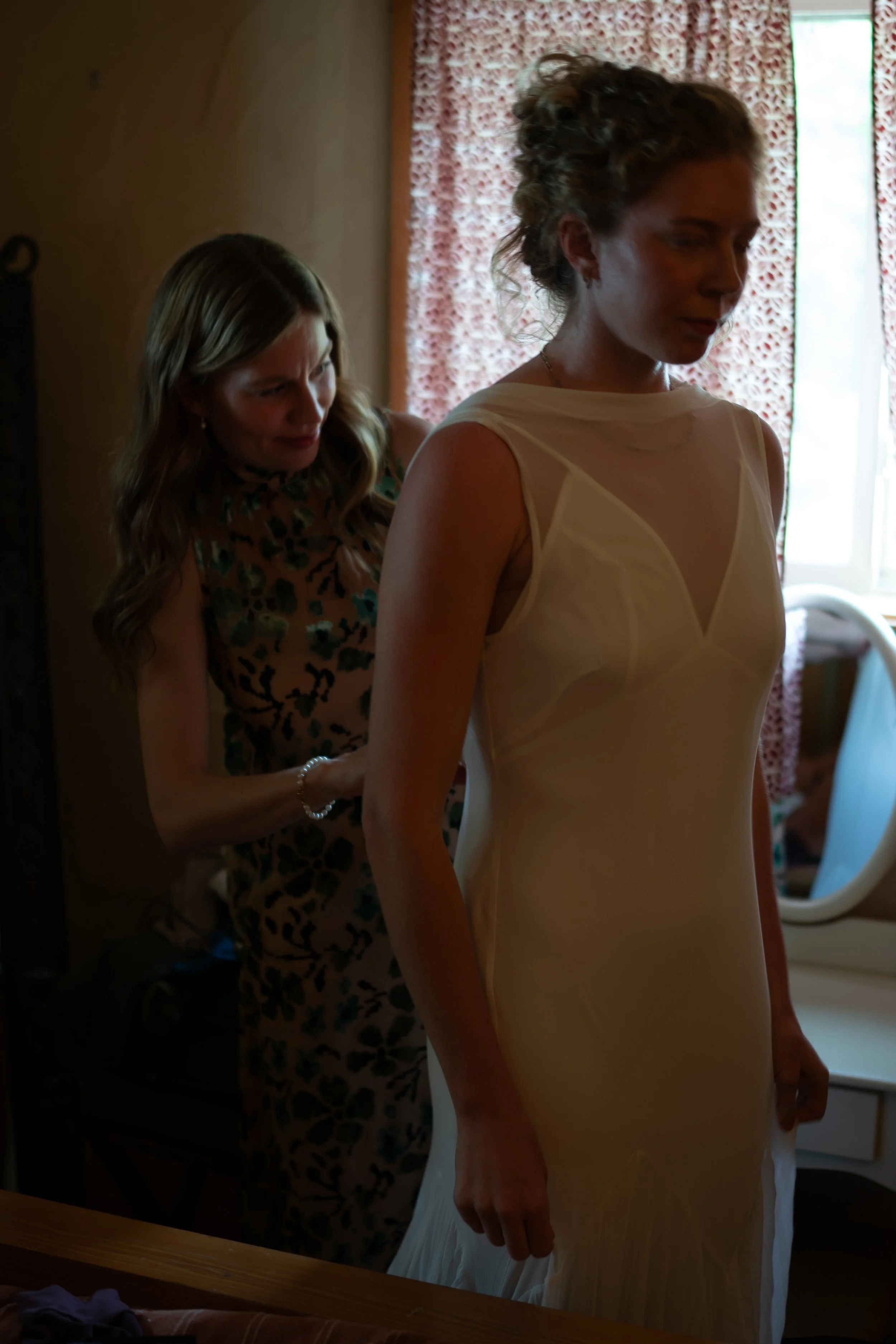 A woman in a white dress is standing while another woman in a leopard print dress is helping her get dressed, possibly for a special occasion, in a warmly lit room with floral curtains.