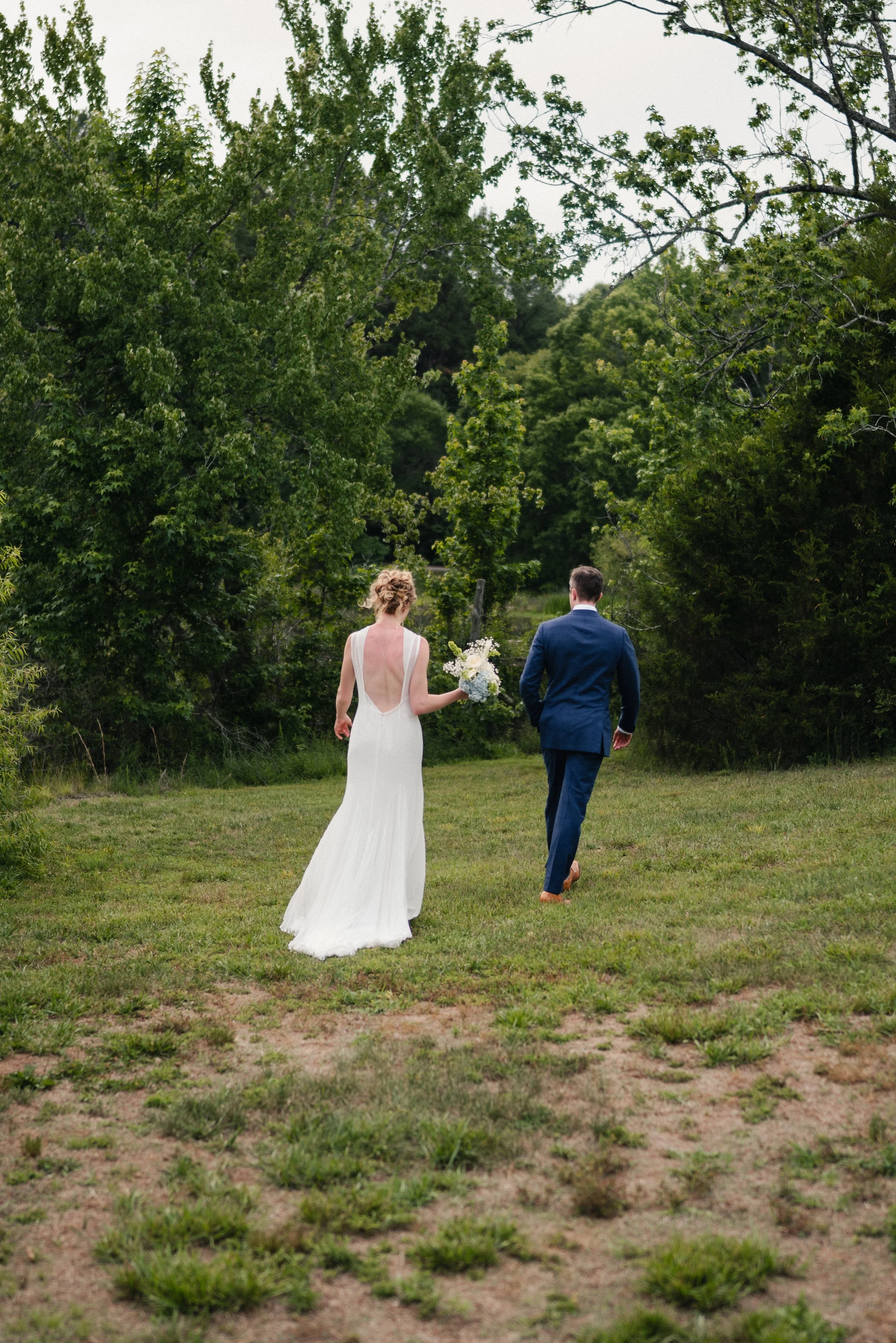 A bride and groom walking away outdoors on a grassy area, surrounded by trees, with the bride holding a bouquet of flowers.