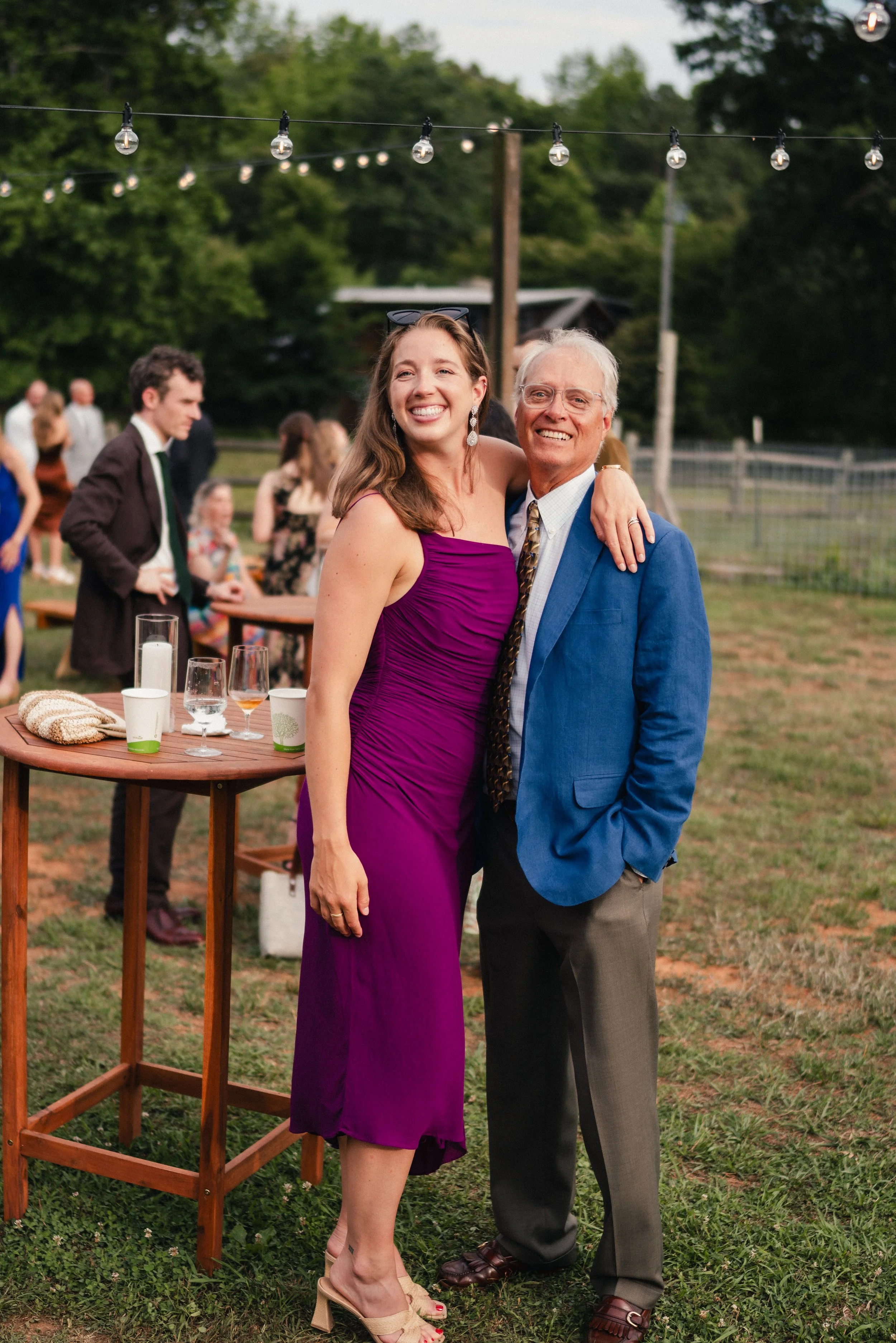 A smiling woman in a purple dress and a smiling older man in a blue blazer pose together outdoors at a celebration, with other guests in the background and string lights above.