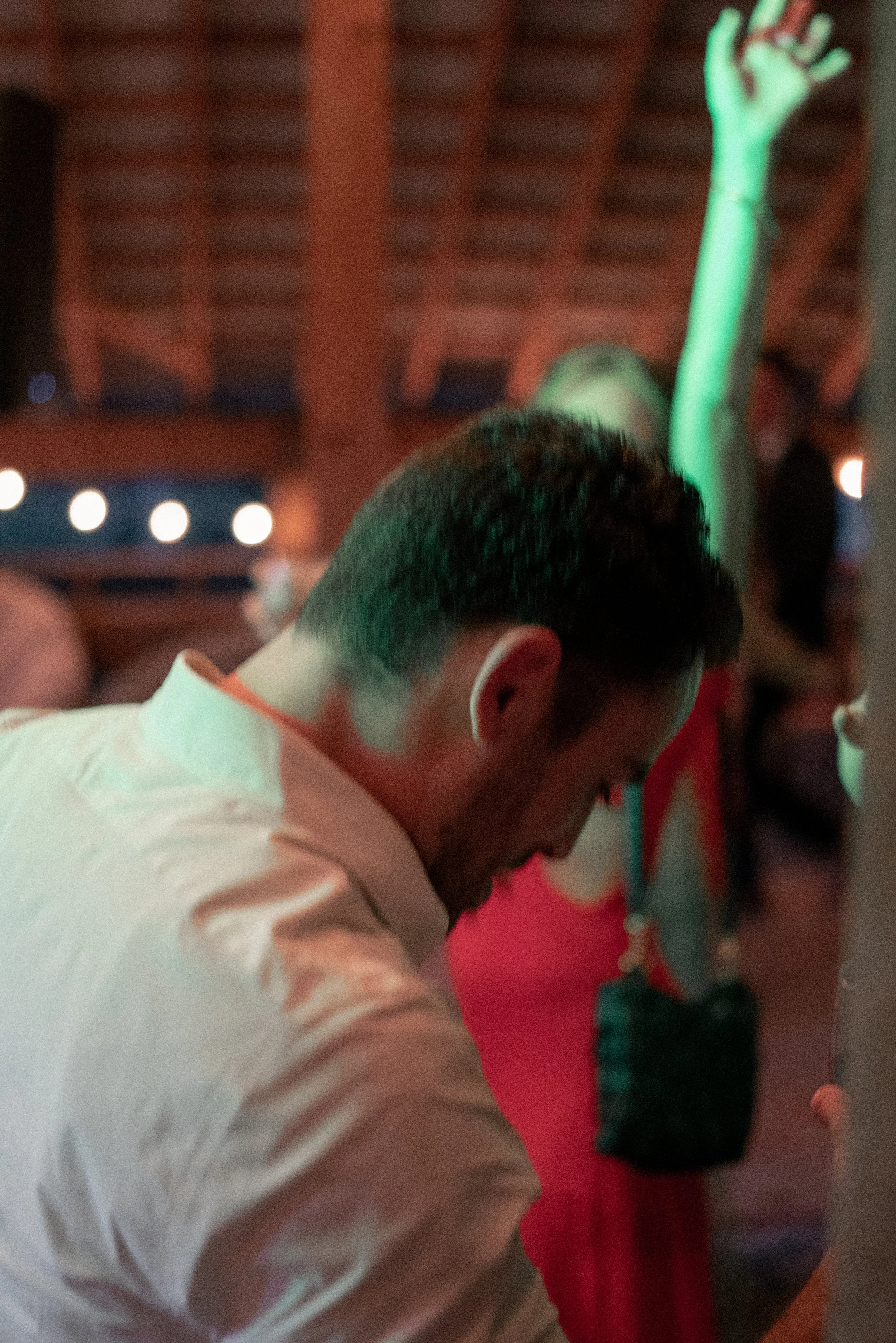 A man with short black hair and a face mask is sitting at a table in a dimly lit environment. His head is bowed, and he appears to be engaged in an activity. Behind him, a woman with long dark hair wearing a red top is standing. The background featur