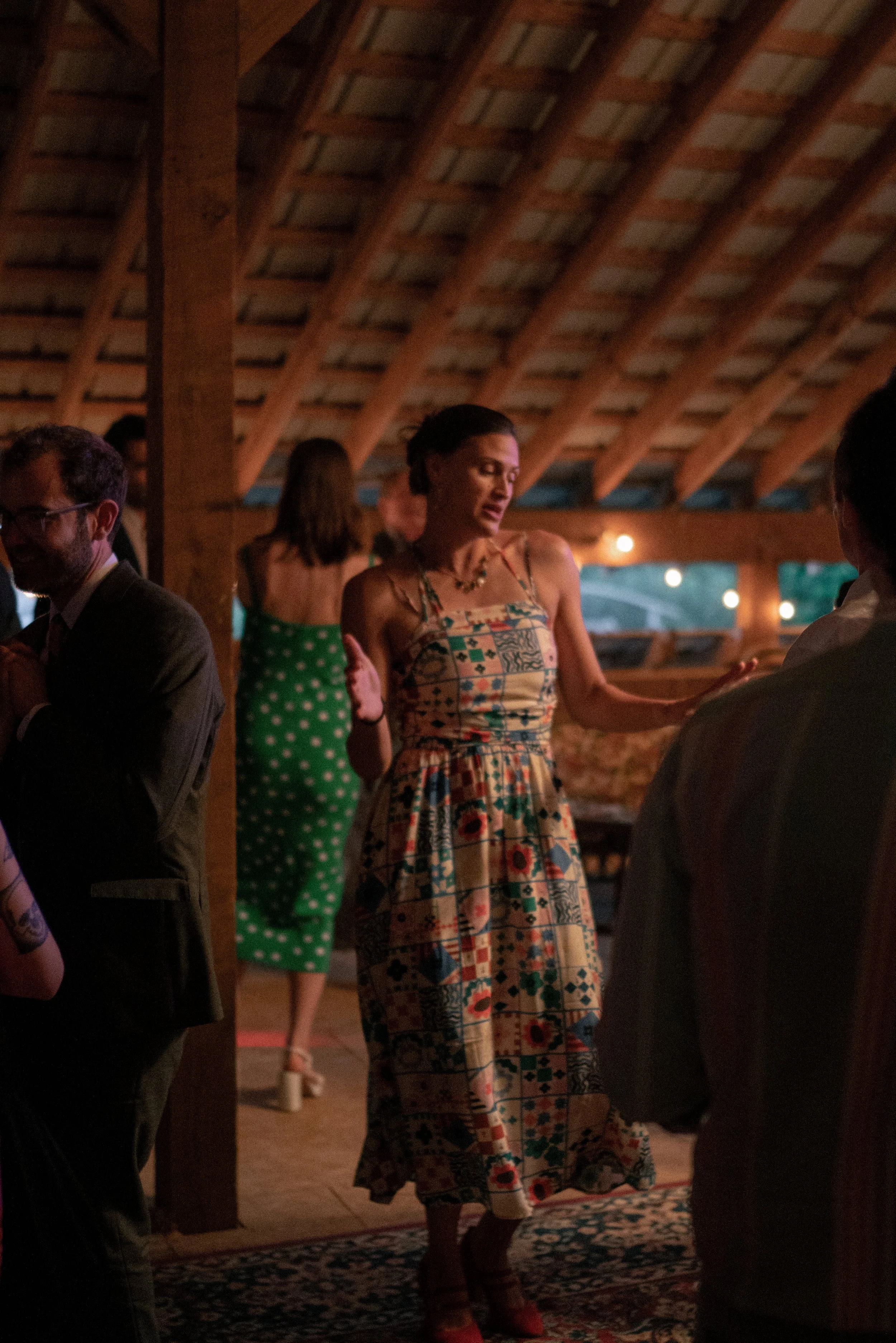 A woman wearing a colorful patchwork dress and red shoes is dancing at an indoor event with warm lighting and a wooden ceiling. Several people are around her, some conversing.