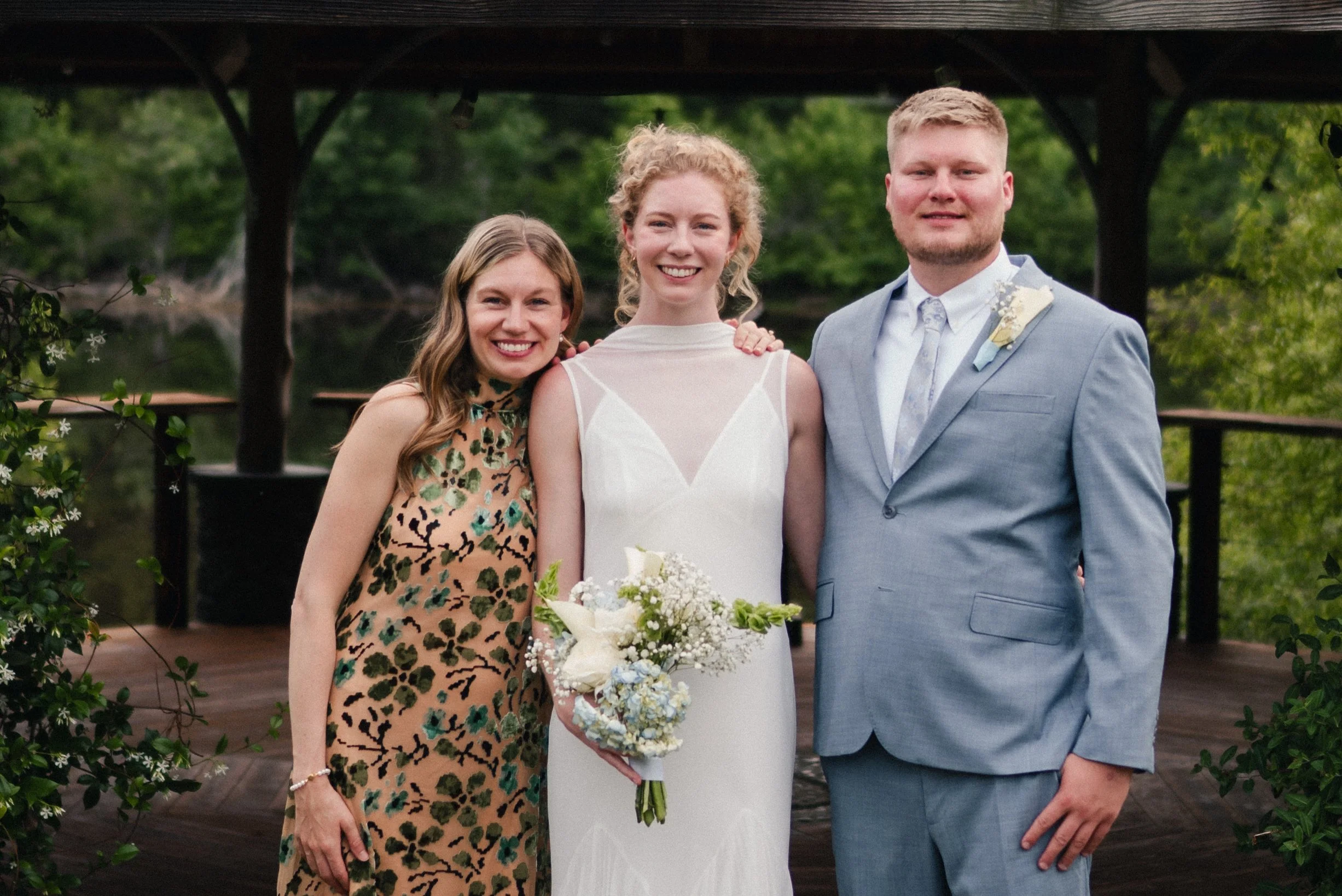 A bride in a white wedding dress holding a bouquet stands between two friends or family members, a woman in a patterned dress on her left and a man in a blue suit on her right, outdoors with greenery in the background.