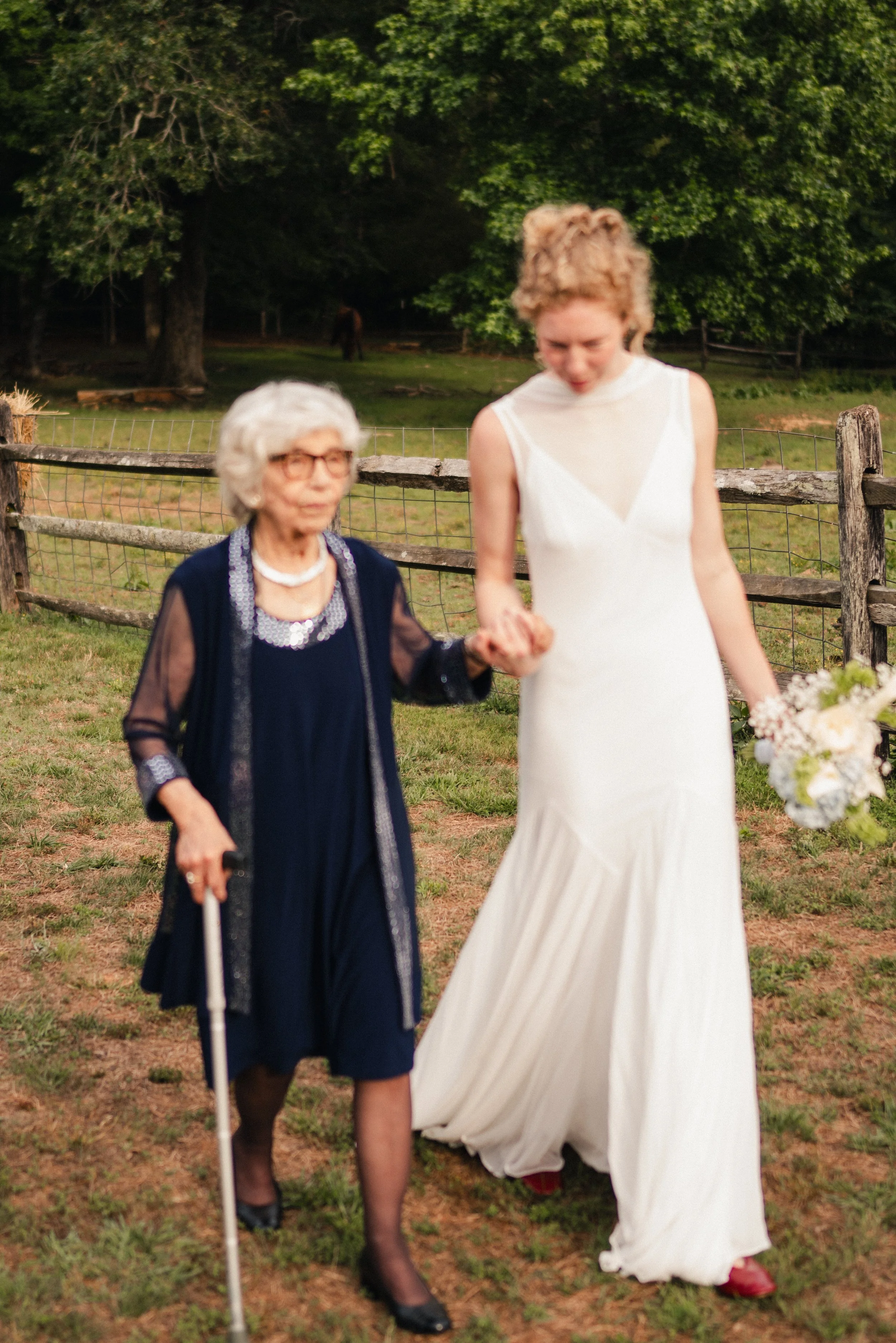 An elderly woman with gray hair and glasses wearing a navy dress with a sheer overlay and a pearl necklace is holding hands and walking with a young woman with short blonde curly hair in a white sleeveless dress outdoors. There is a wooden fence and 
