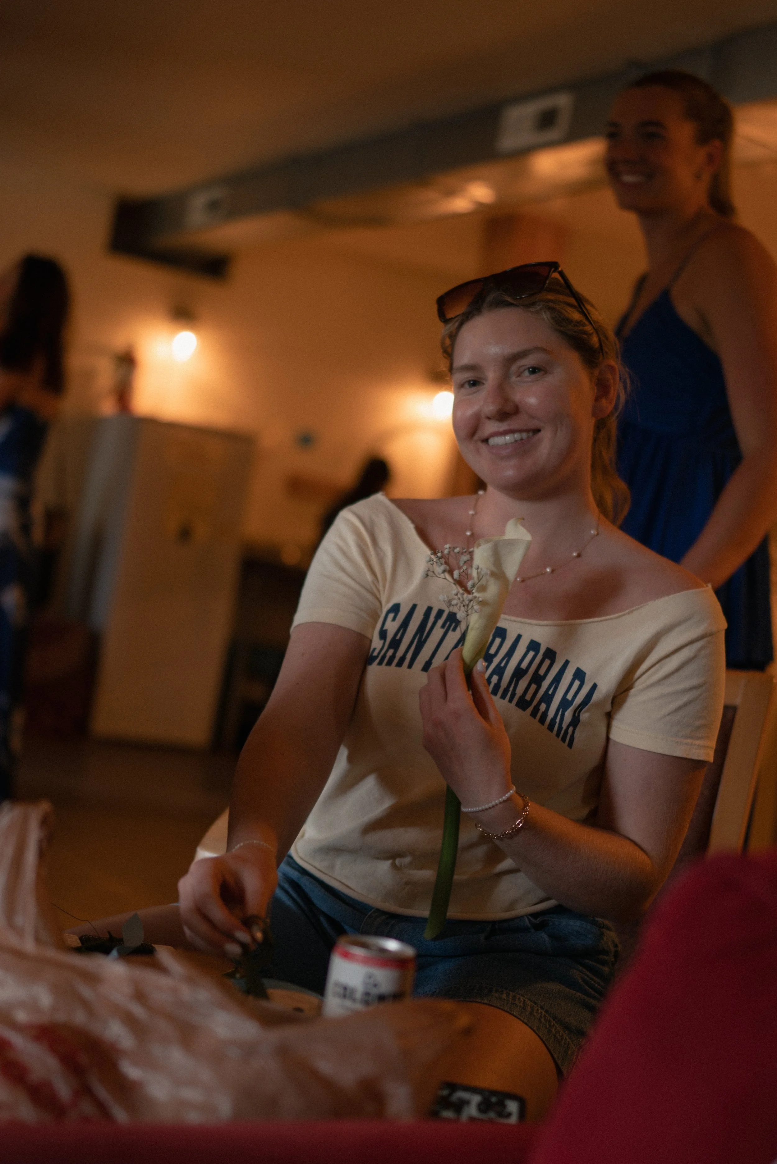 Smiling young woman with glasses on her head, holding a flower, wearing a beige T-shirt with 'SANTA BARBARA' written on it, sitting in a cozy indoor setting, with another woman smiling in the background.