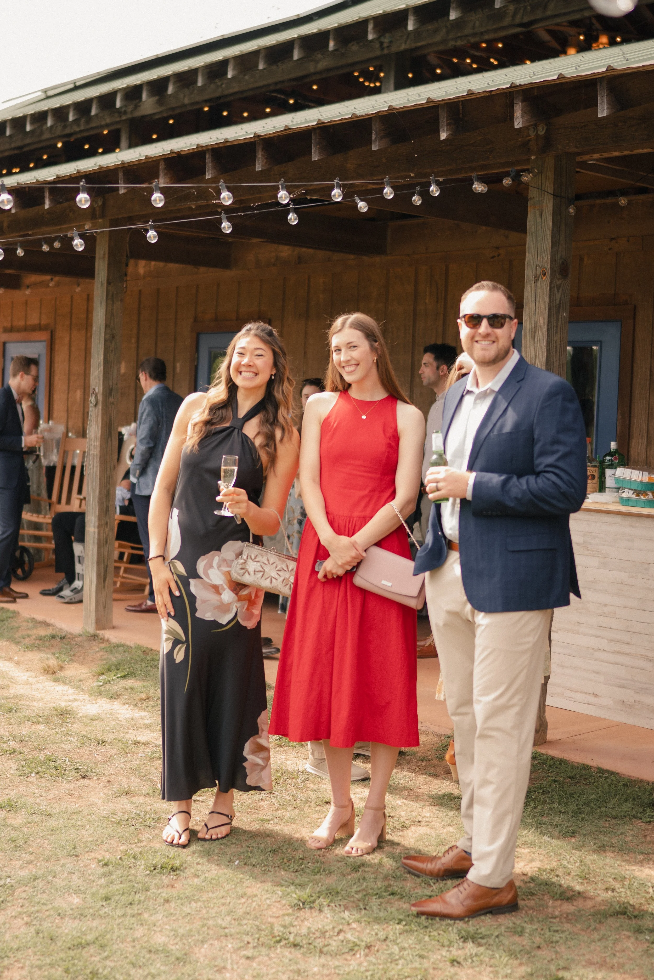 Group of four young adults, two women and two men, smiling and dressed in semi-formal attire at an outdoor event held in a rustic, wooden venue with string lights overhead. The women are holding glasses of champagne, and the man is holding a bottle. 