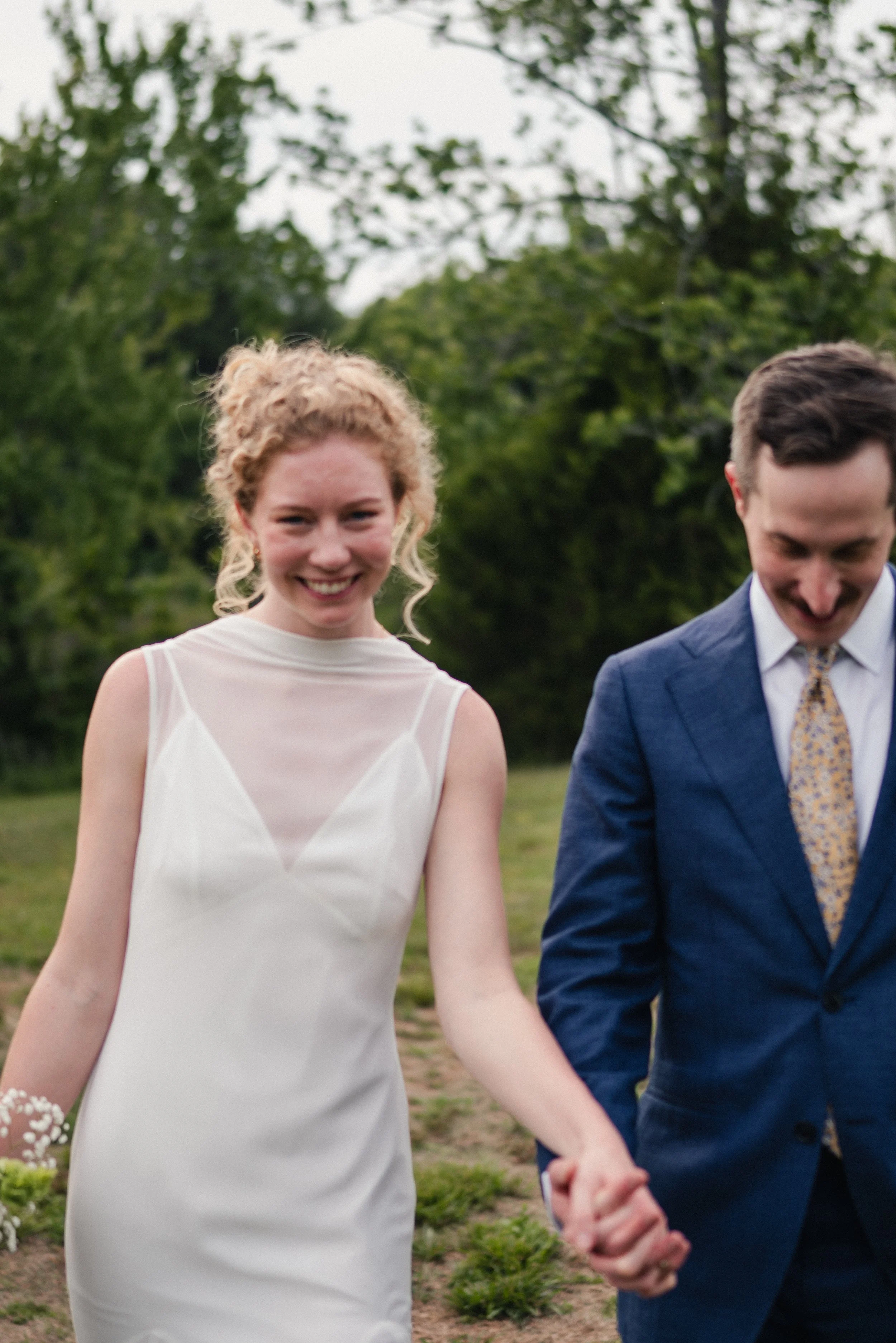 A smiling woman with curly blonde hair wearing a white wedding dress holding hands with a man in a blue suit, outdoors with green trees in the background.