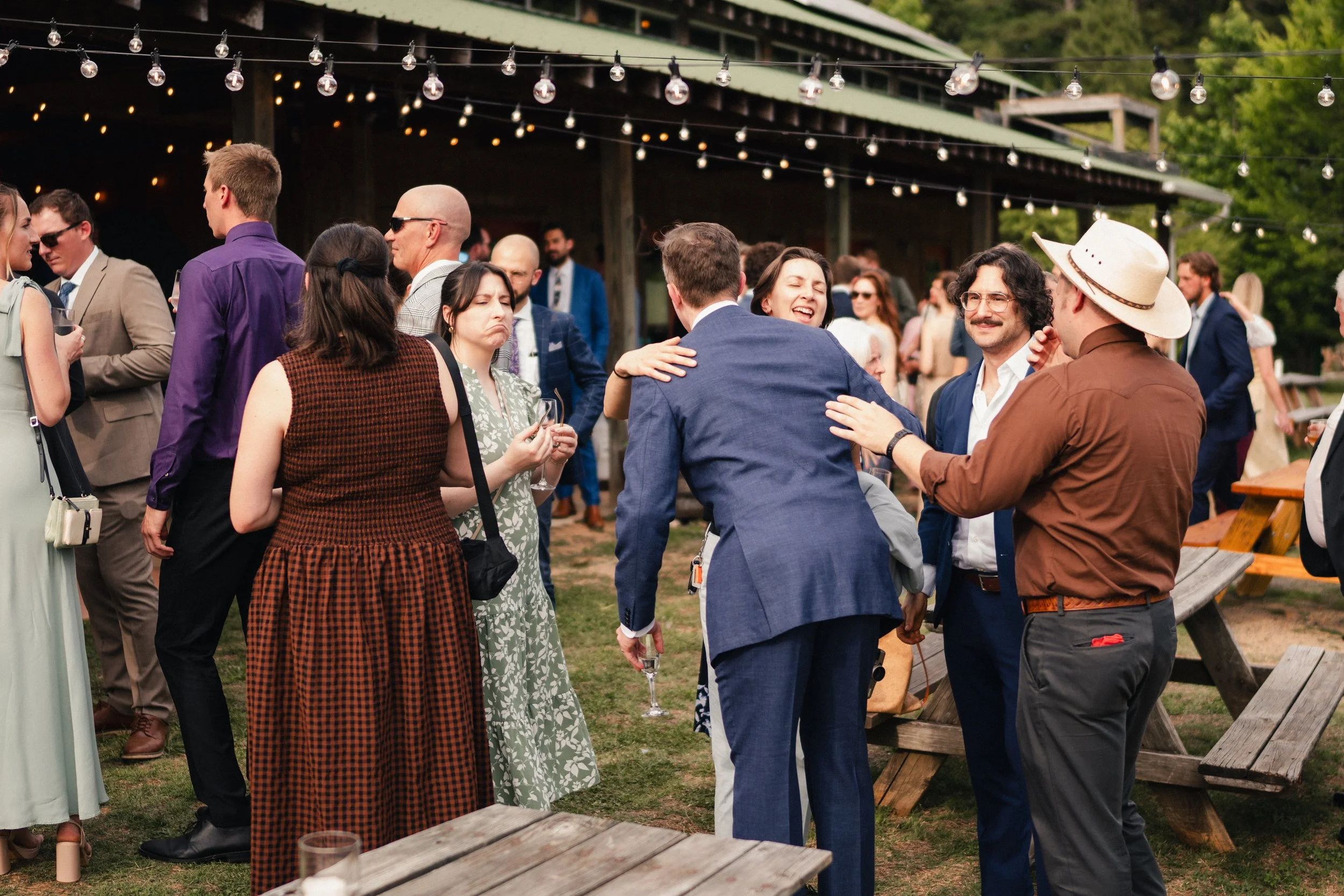 A group of people socializing outdoors at a party, some embracing, some holding drinks, with string lights hanging overhead and a wooden building in the background.