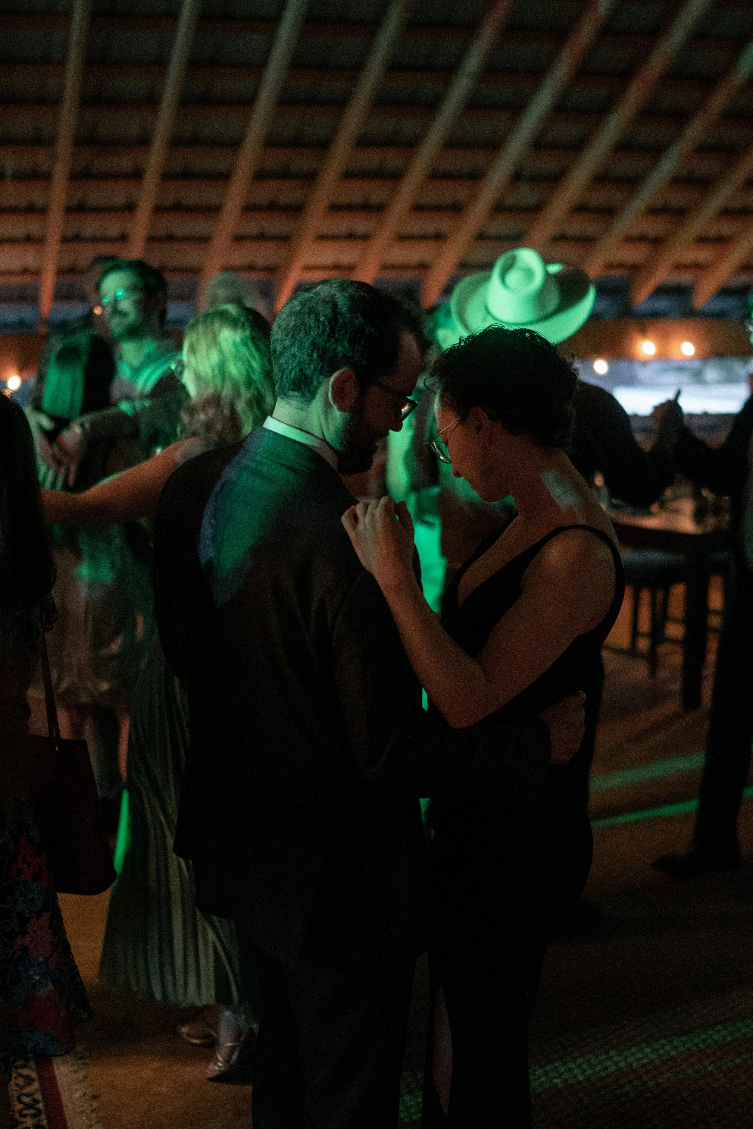 A couple dancing intimately in a dimly lit, rustic venue with a wooden ceiling and warm string lights in the background, surrounded by other people enjoying the event.