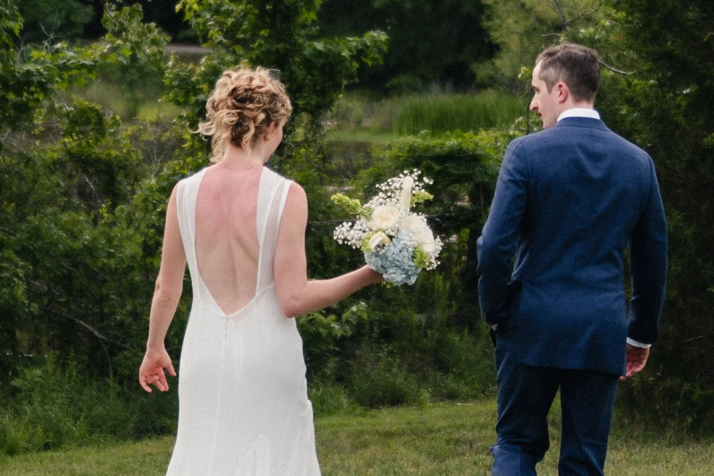 A bride in a white wedding dress with an open back holds a bouquet of white and green flowers while walking with a groom in a blue suit outdoors among greenery.