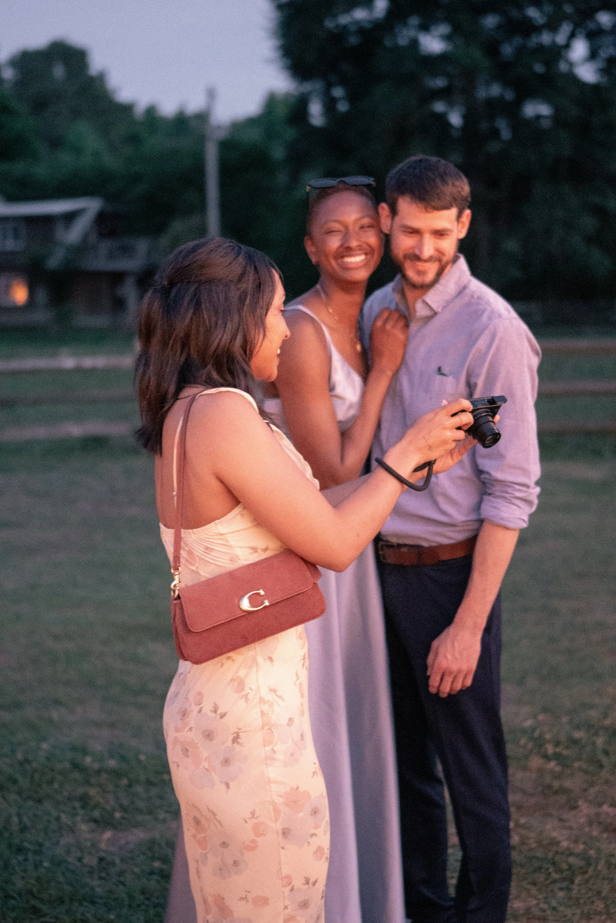 Four friends are gathered outdoors at dusk, taking a selfie together. They are smiling and appear happy, with a background of trees and a house with lights on.