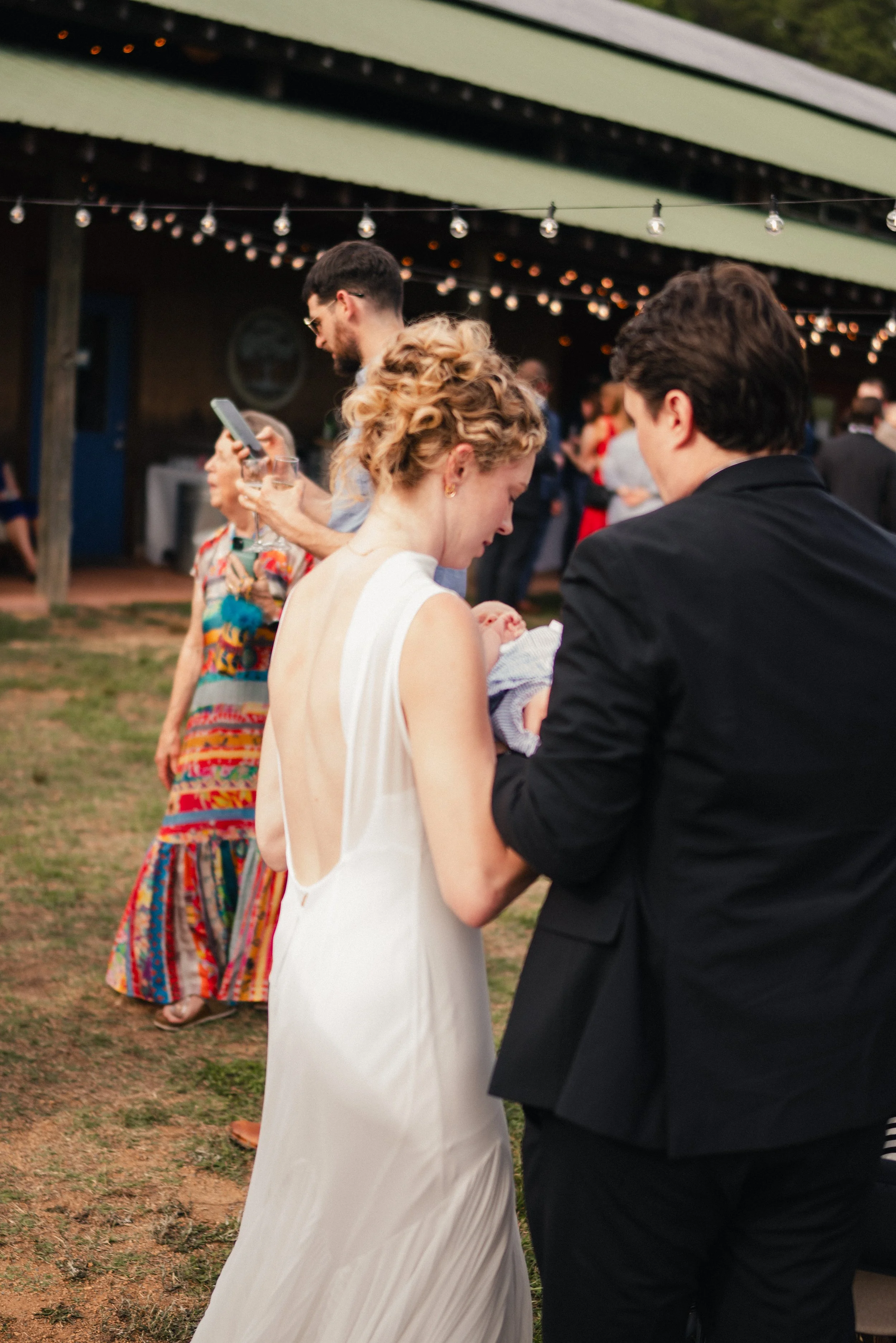 A group of people at an outdoor gathering under string lights, with a woman in a white dress holding a baby, and a man in a black suit standing next to her. In the background, there is an older woman in a colorful dress.