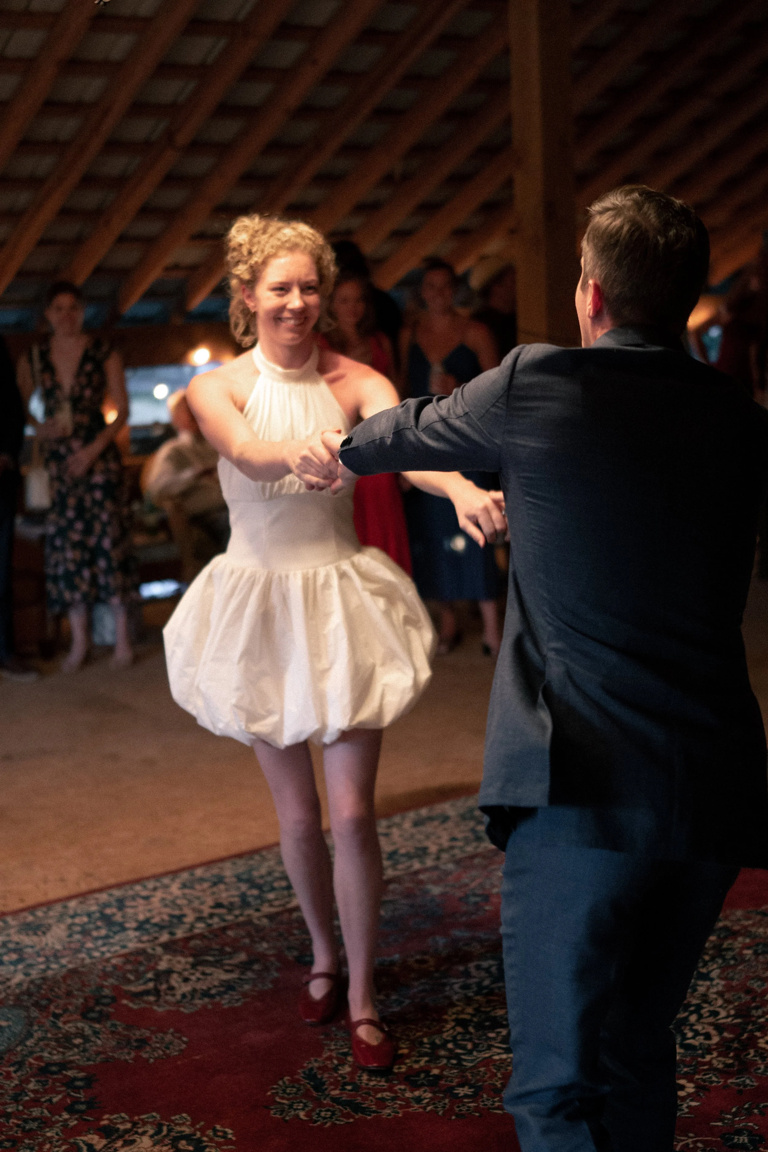 A woman in a white dress with a puffed skirt is dancing with a man in a dark suit at an indoor gathering. Other people are watching and socializing in the background.
