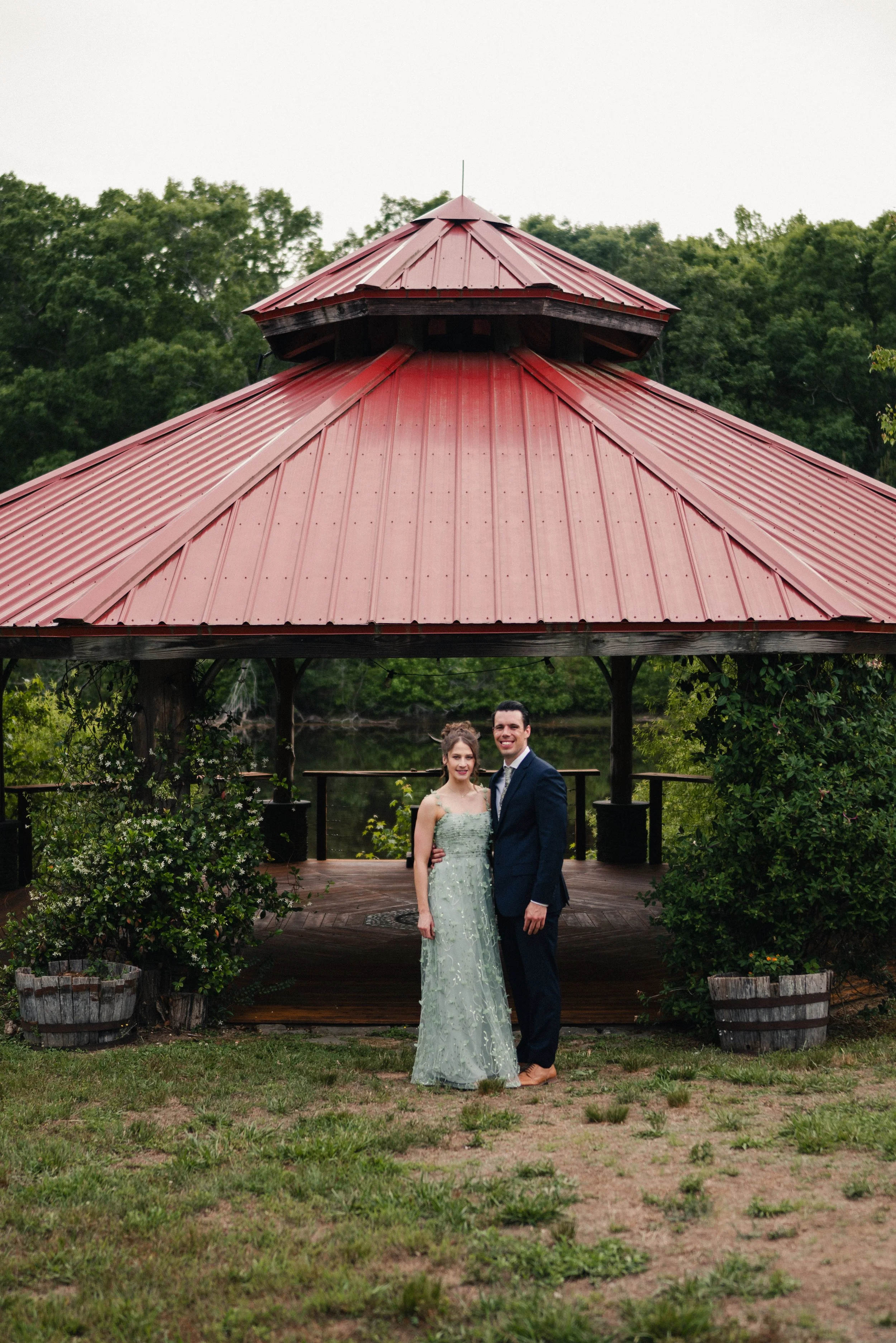 A couple dressed in wedding attire standing in front of an open-air pavilion with a red metal roof, surrounded by greenery and trees, near a body of water.