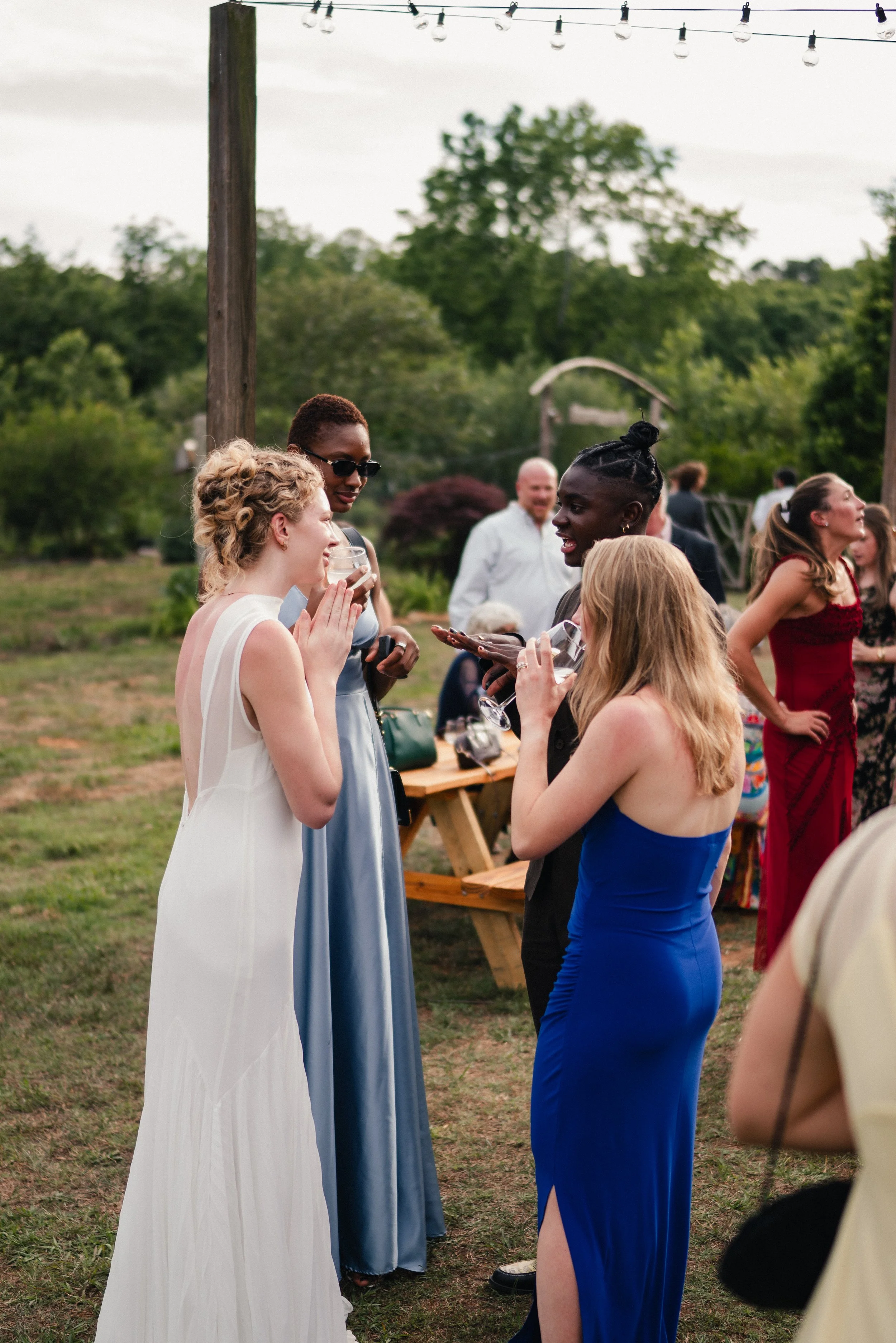 A group of women in elegant dresses are engaging in conversation at an outdoor event, with string lights overhead and greenery in the background.