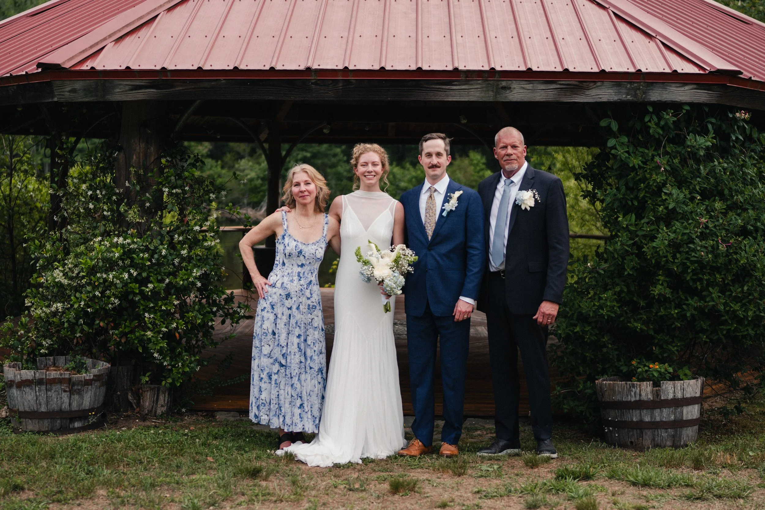 Group of four people standing outdoors, two women and two men, dressed in wedding attire, posing for a photo in front of a wooden pavilion with a red metal roof, surrounded by greenery.