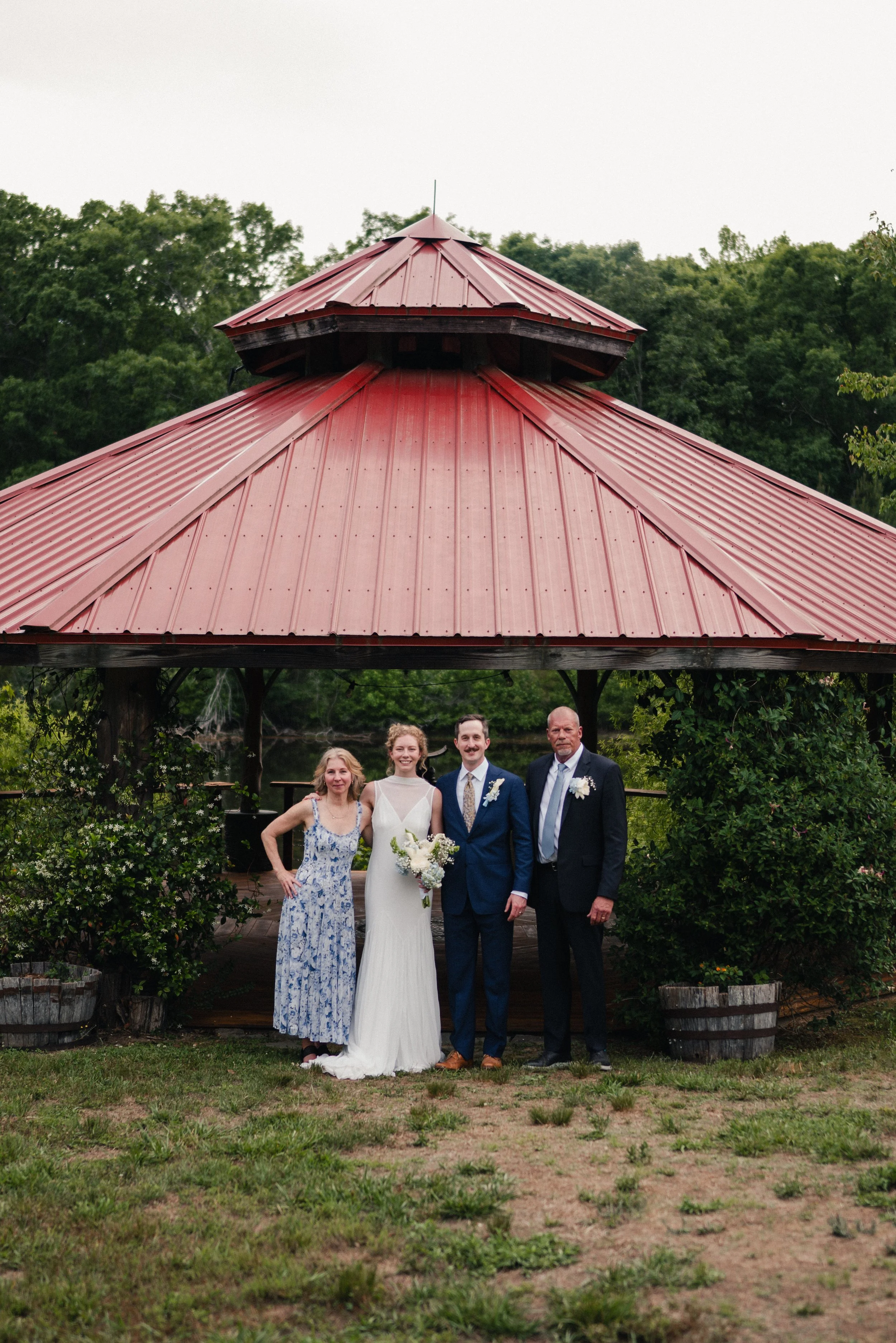 Four people pose for a photo in front of a red pavilion with a metal roof, surrounded by greenery and trees, during daylight.