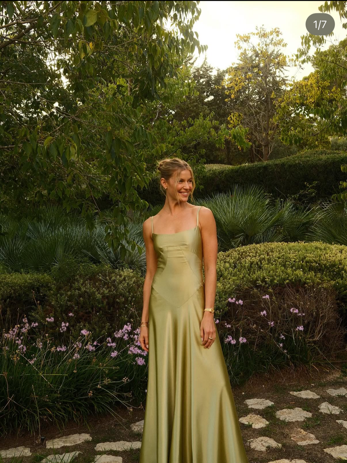 A woman in a light green satin dress is standing outdoors in a garden with lush green trees, bushes, and purple flowers, smiling and looking to the side.