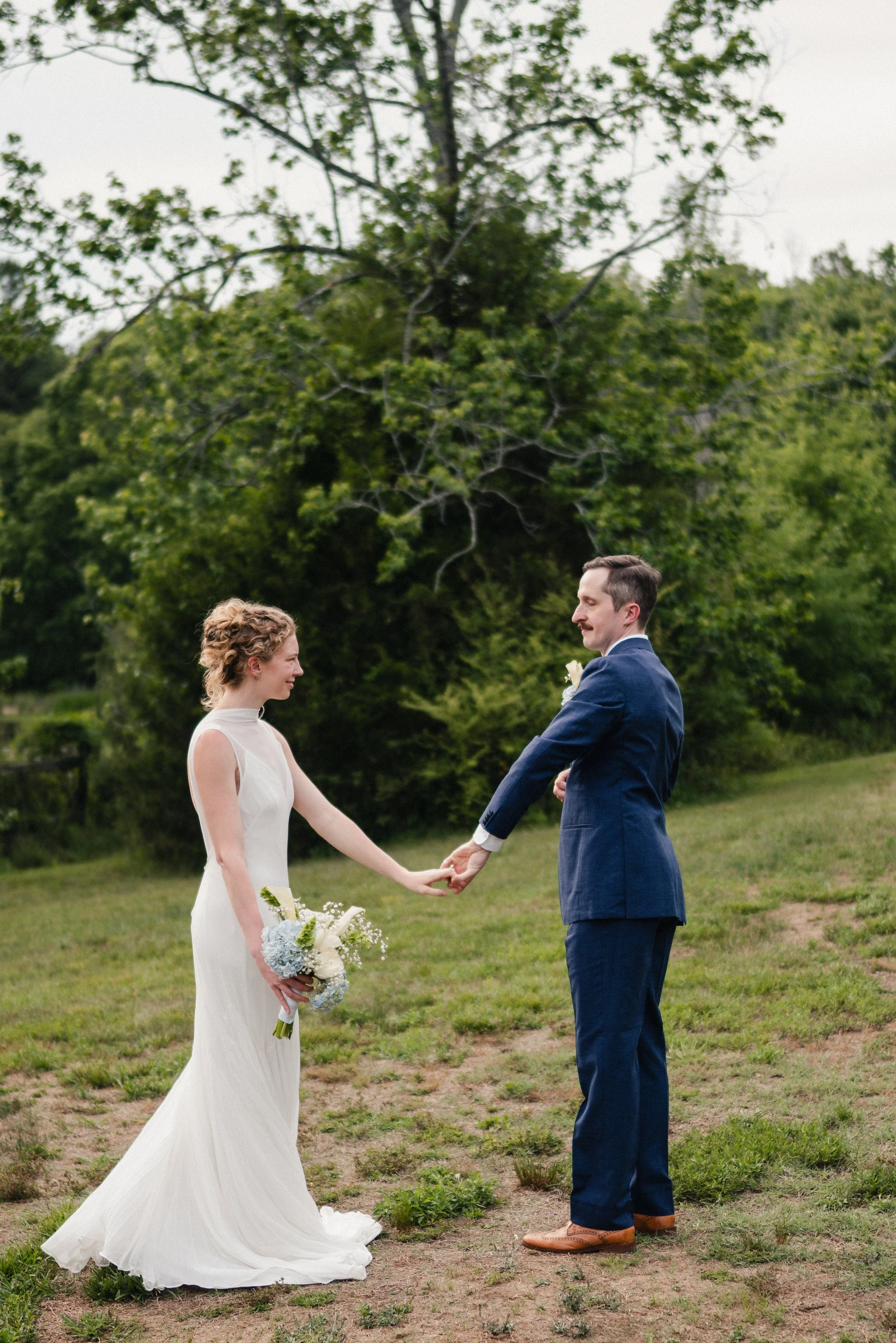 A bride and groom standing outdoors holding hands and looking at each other during their wedding ceremony in a lush green area with trees in the background.