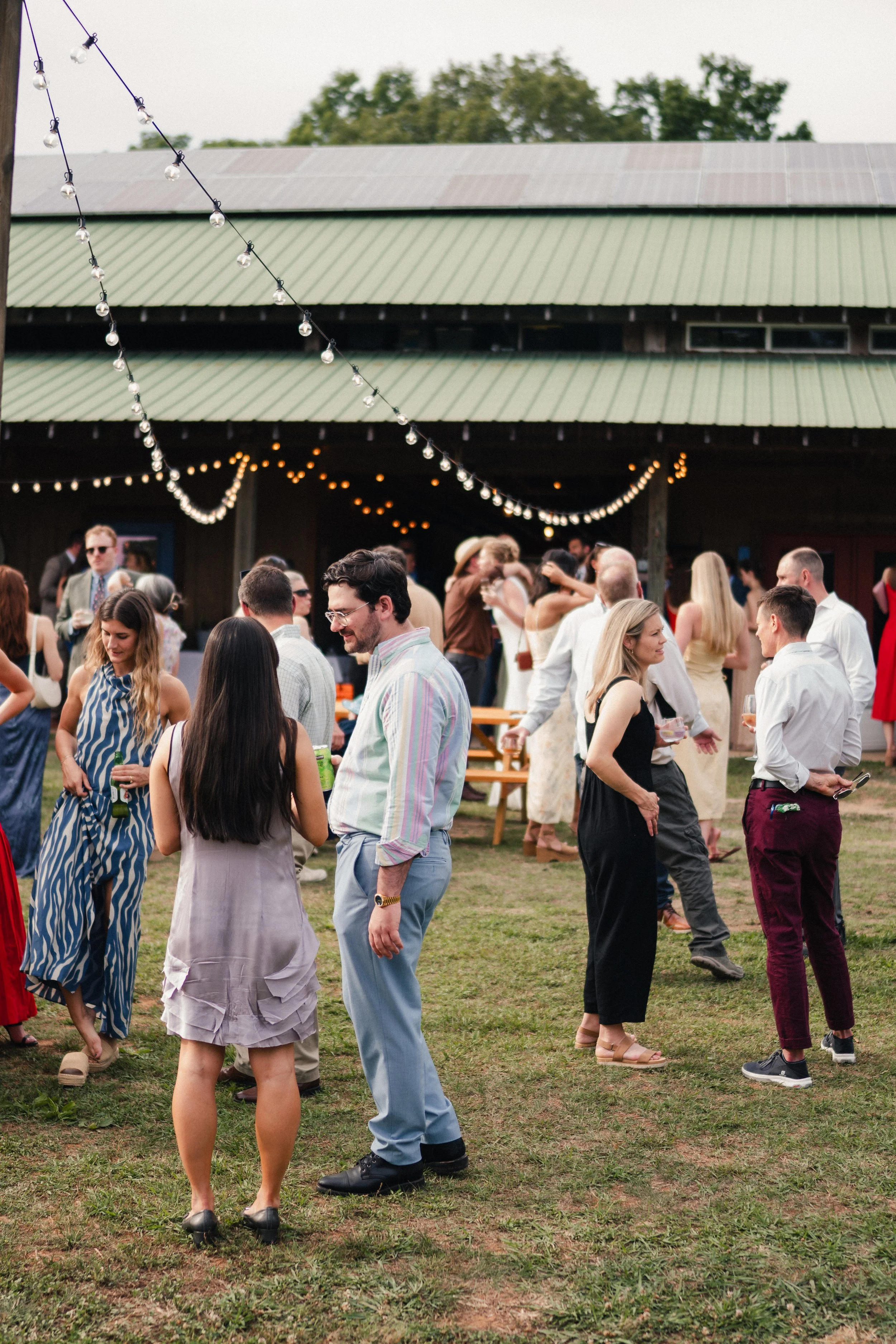 People socializing outdoors at a gathering under string lights near a barn with a green metal roof.