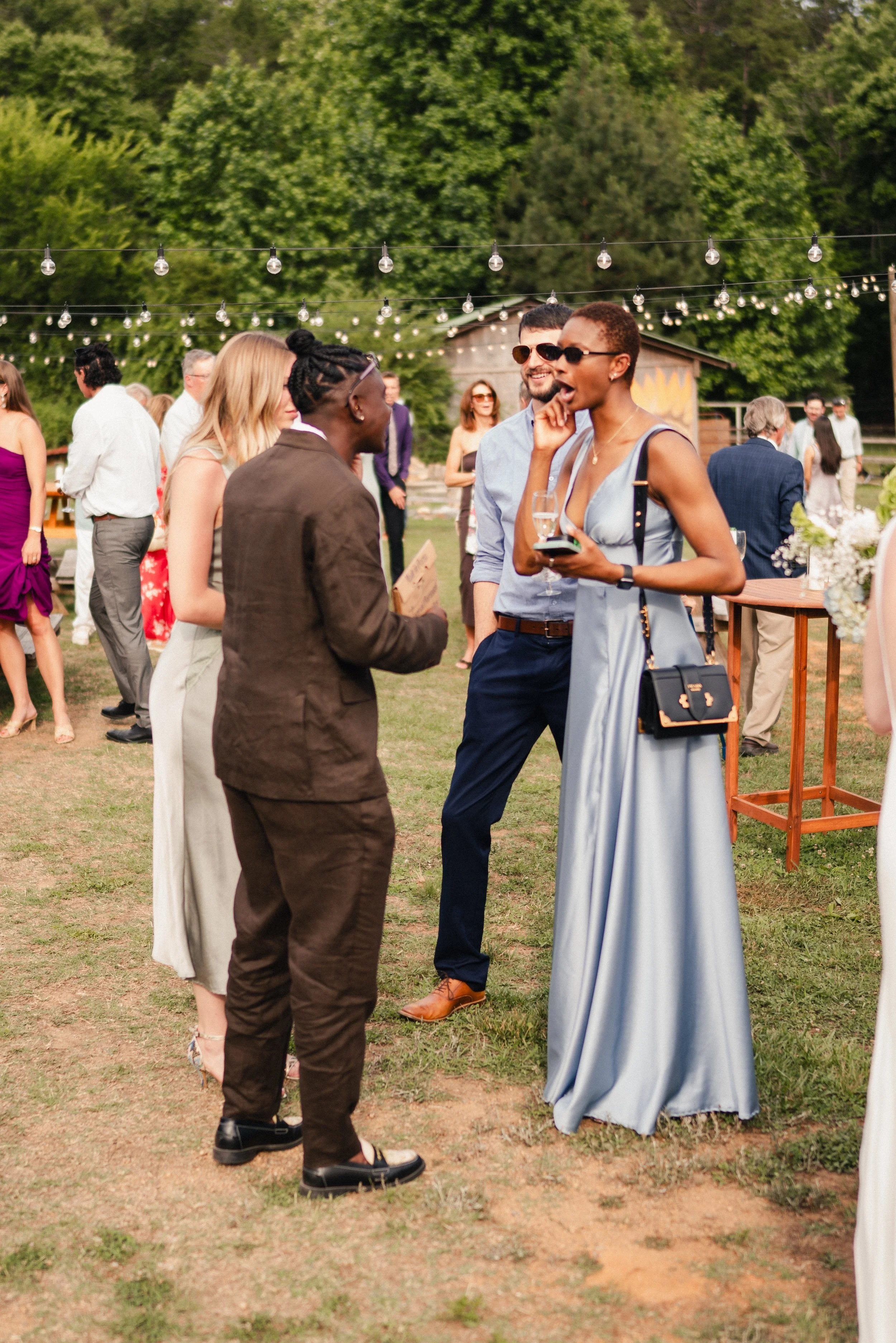 A group of people socializing outdoors at a party with string lights overhead, greenery in the background, and a relaxed, festive atmosphere.