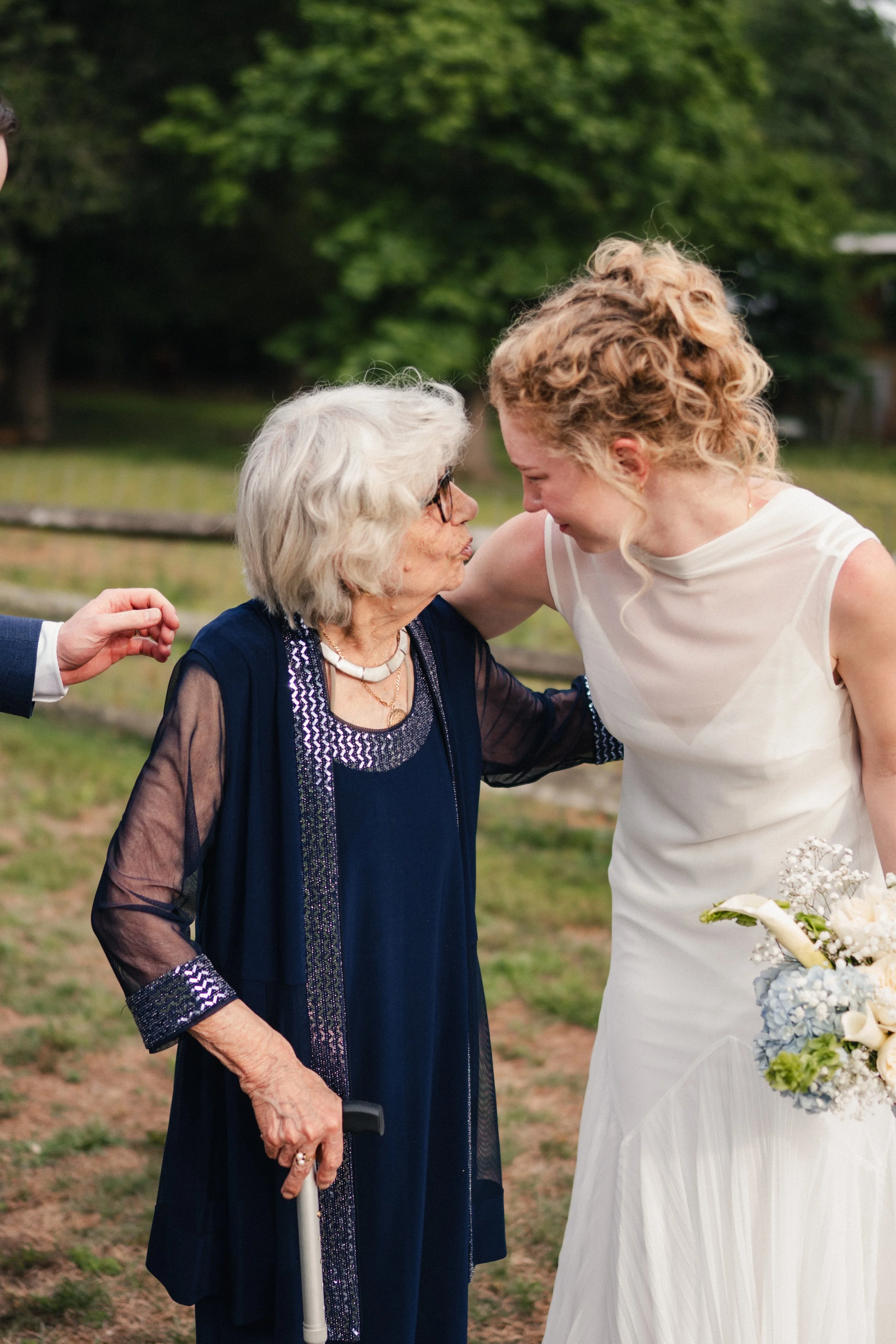 A young woman in a white wedding dress is leaning her forehead against an elderly woman, who is dressed in a navy blue dress with intricate beading and is holding a white cane. They are outdoors, with trees and grass in the background, and appear to 