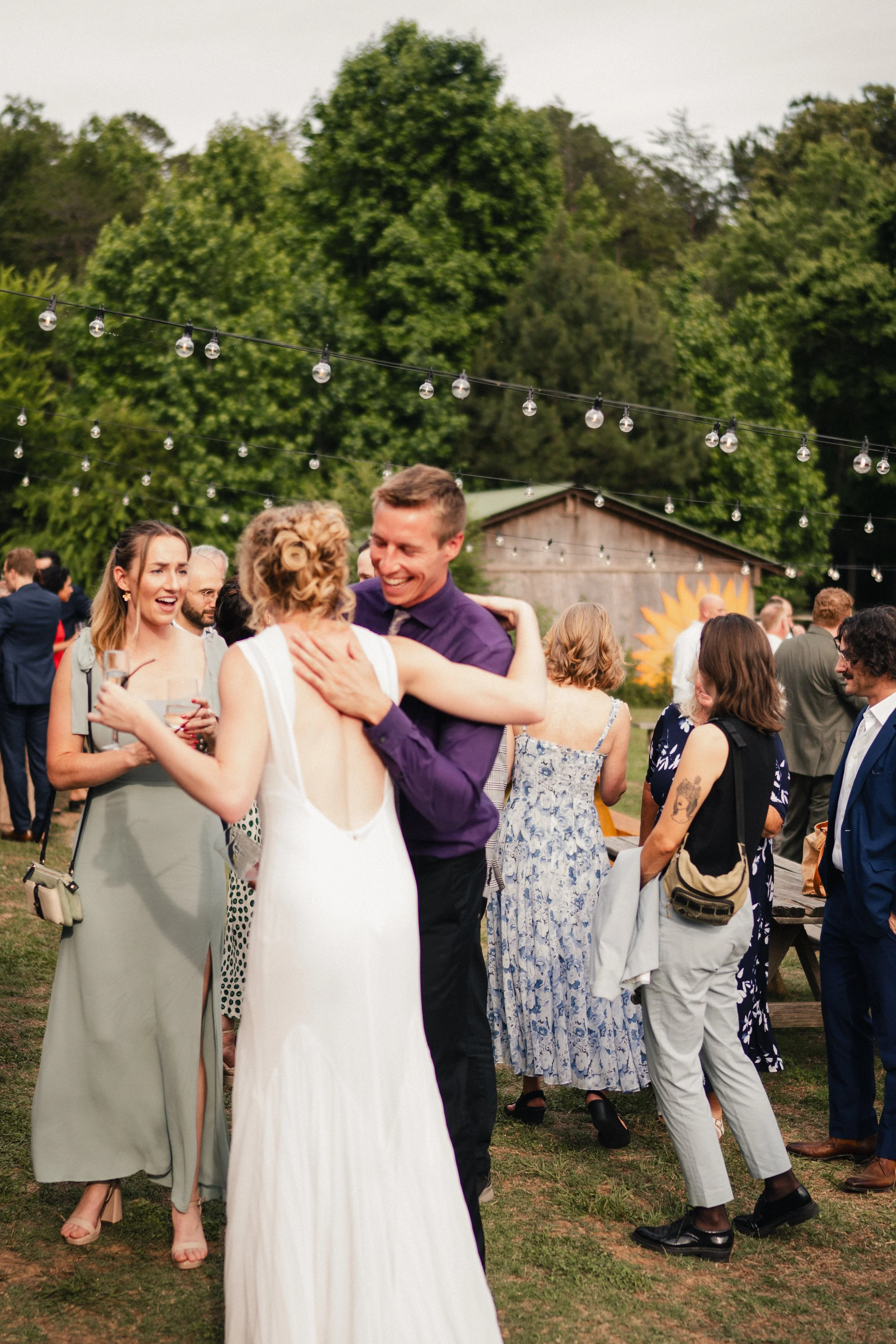 People dancing and socializing outdoors at a celebration or event, with string lights overhead and trees in the background.