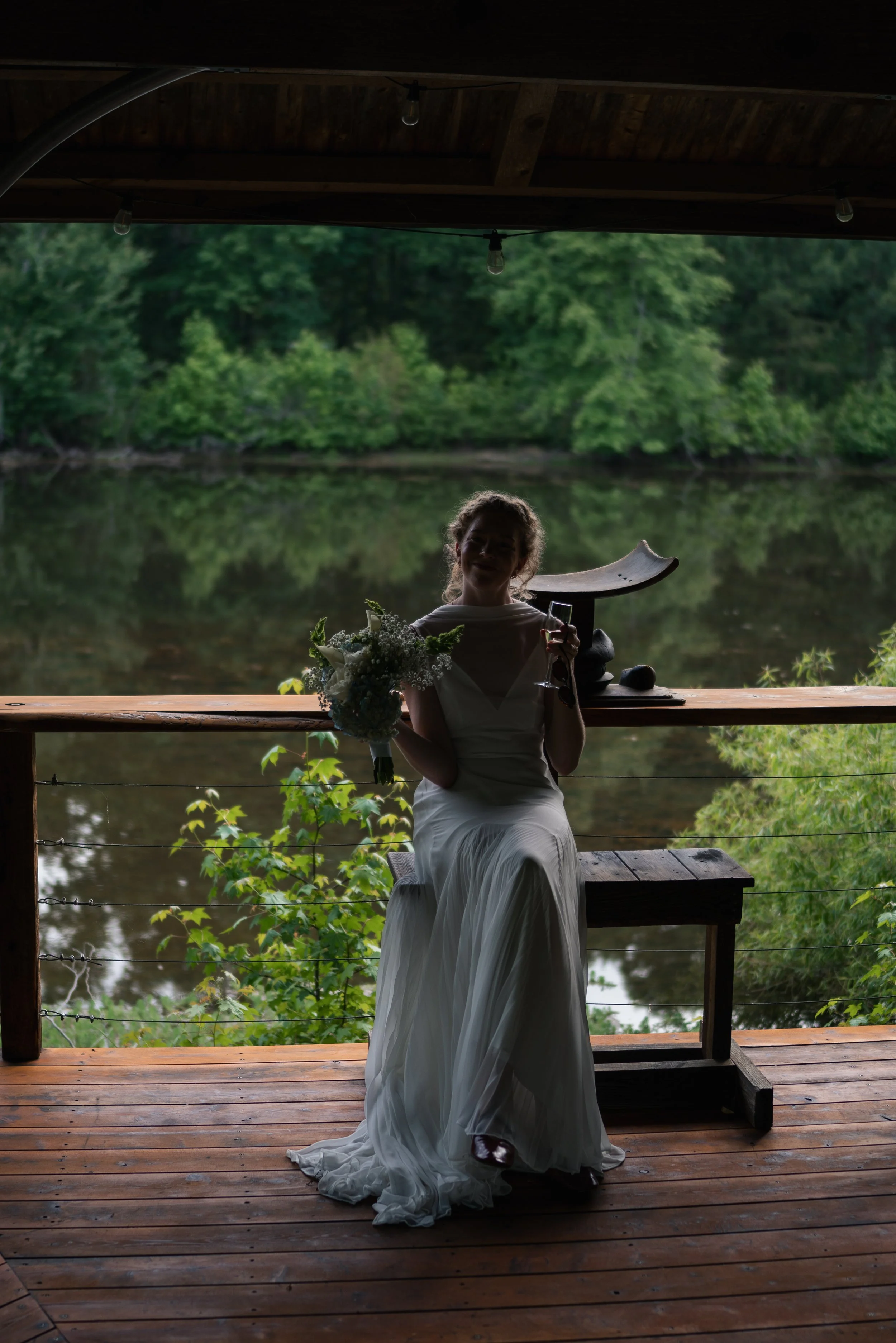 A woman in a white dress sitting on a bench on a wooden deck near a body of water, holding a bouquet of flowers and a glass of wine, with lush green trees in the background.
