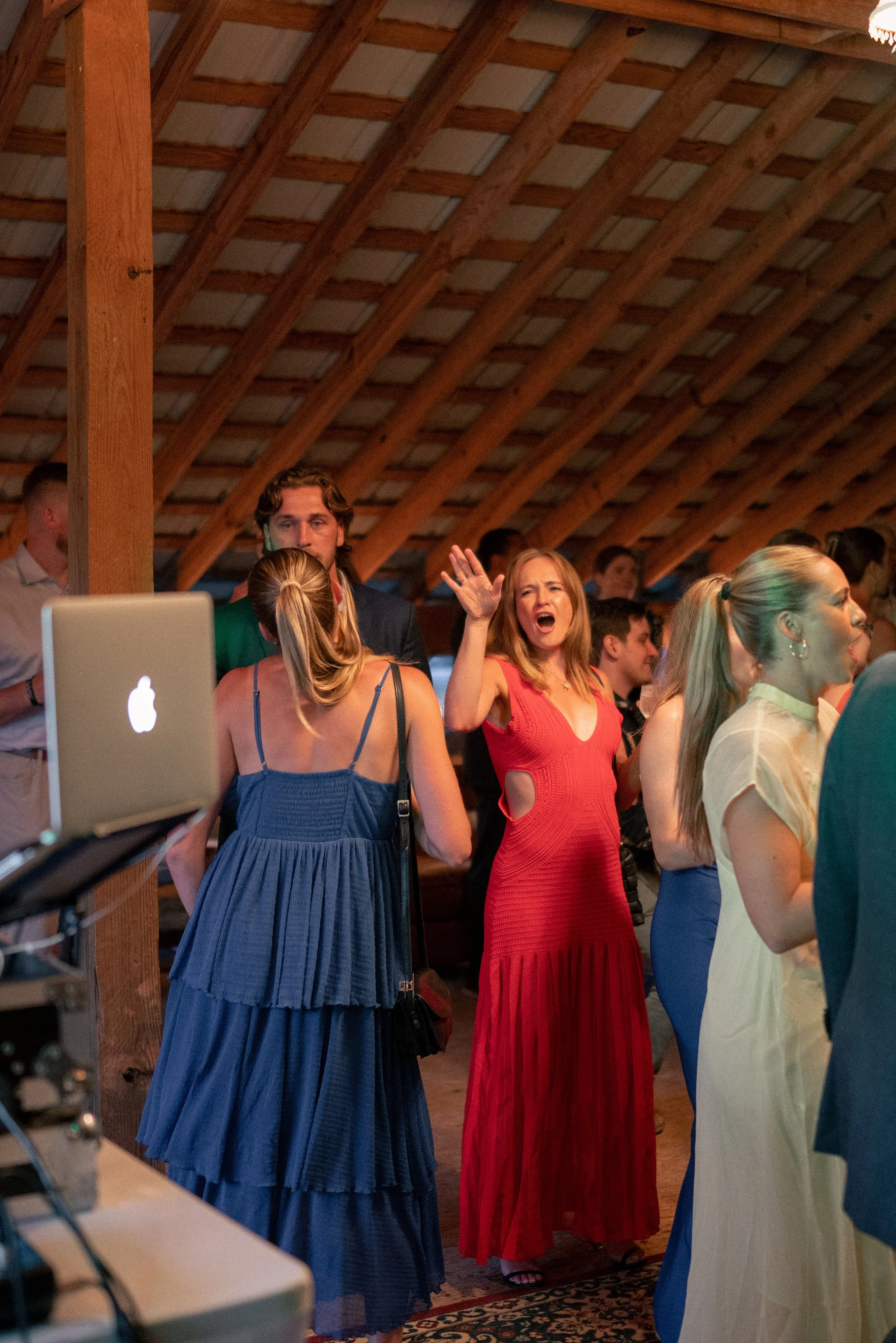 People gathered at an indoor event with wooden ceiling beams, including a woman in a red dress raising her hand and talking, surrounded by others in various colorful dresses.