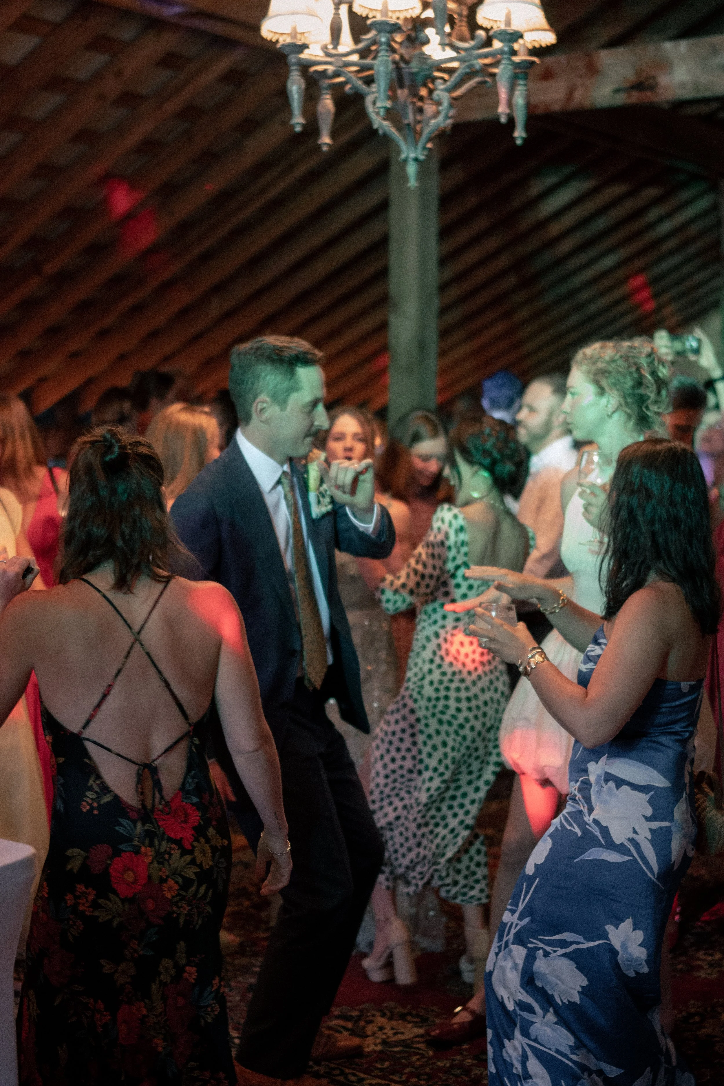 A group of people dancing at a party or wedding reception in a dimly lit room with wooden beams. The focus is on a man in a suit and a woman in a blue floral dress, with other guests in the background.