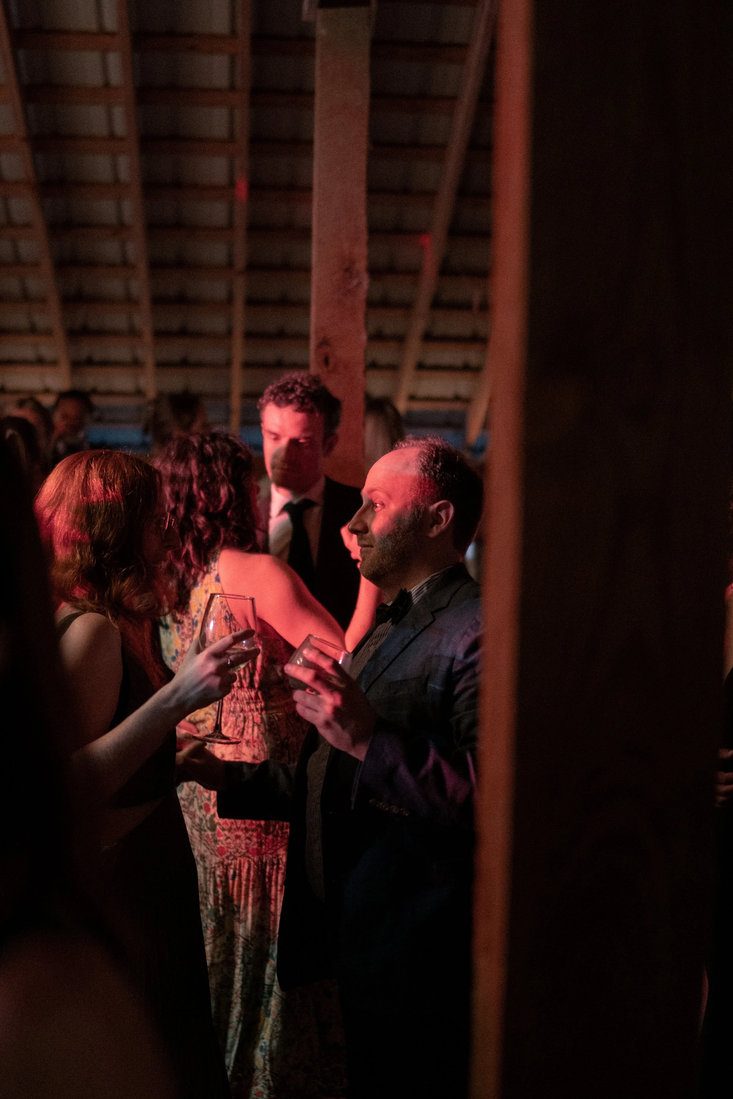 A group of people dressed in formal attire socializing at an indoor event, with some holding wine glasses, in a dimly lit setting with wooden beams on the ceiling.
