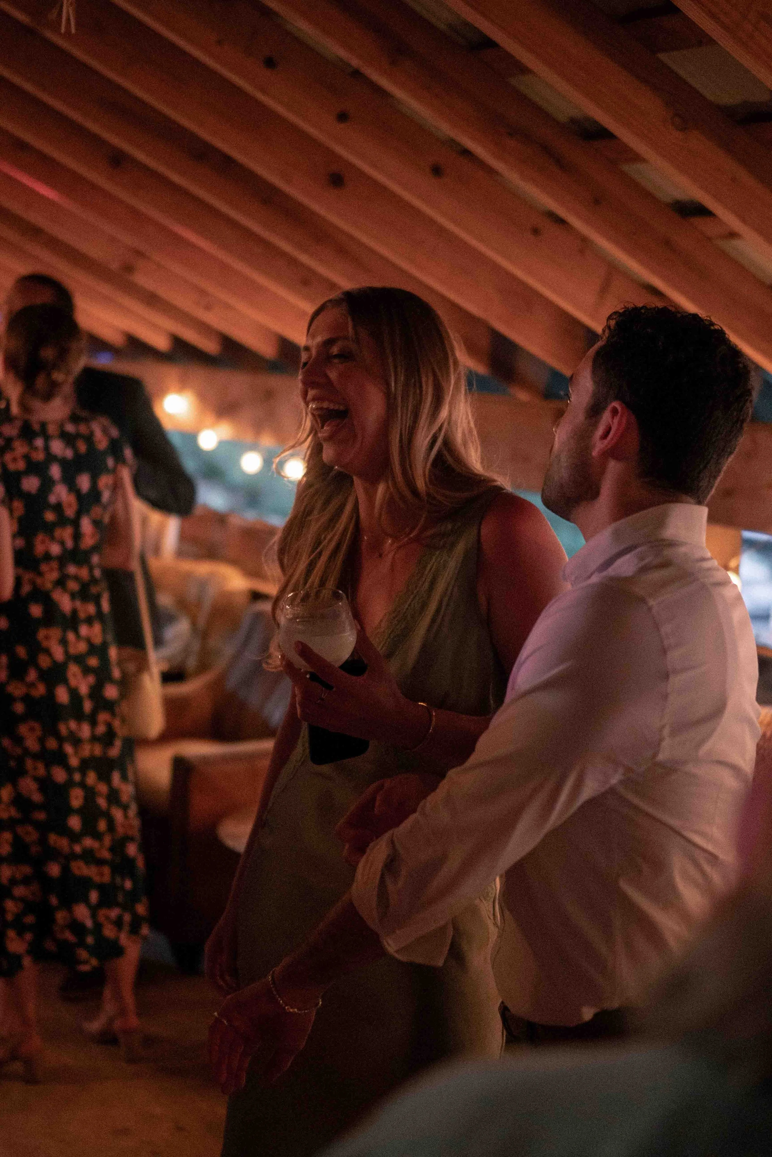 A woman and a man are laughing and talking at a social gathering in a warmly lit room. The woman holds a drink and has long, wavy hair. The man wears a white shirt and has short, dark hair. Other guests are visible in the background.
