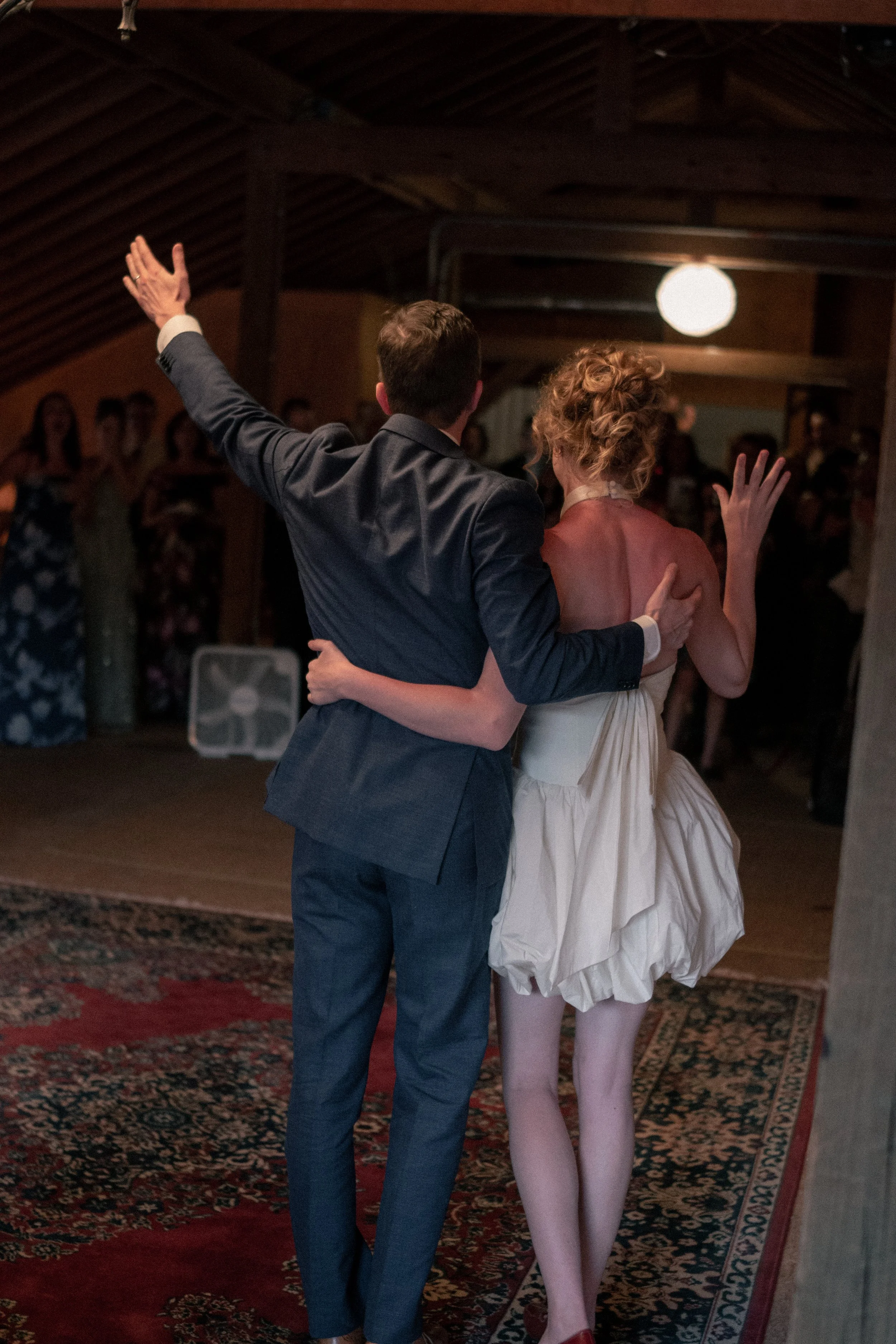 A man and a woman are dancing together in a dimly lit room with wooden ceiling beams and a patterned rug. The man is wearing a gray suit, and the woman is wearing a beige dress. The man has his arm raised, and the woman has her arm around his shoulde