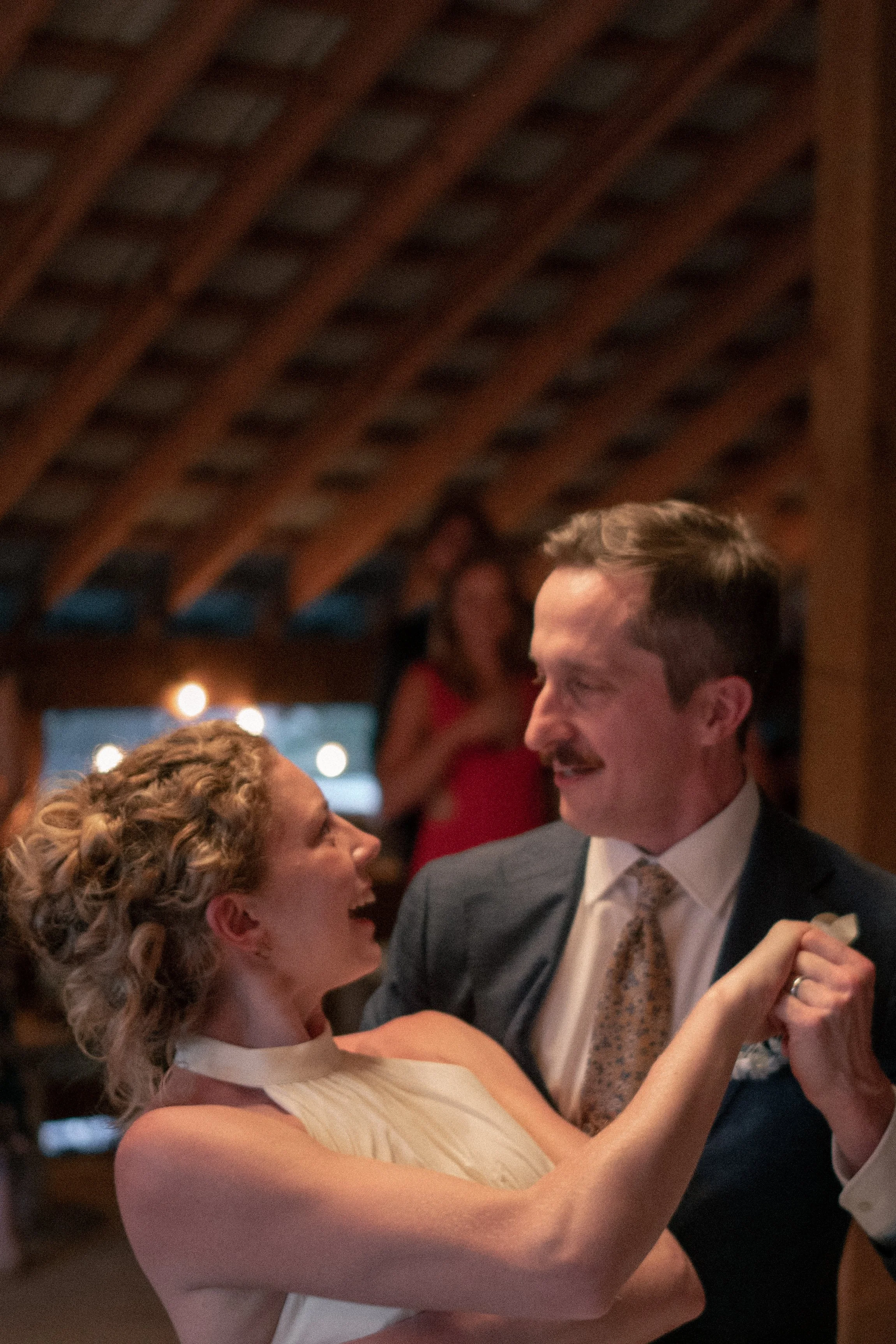 A woman and a man are dancing closely at a social event, smiling and looking at each other, with warm indoor lighting and wooden ceiling beams overhead.