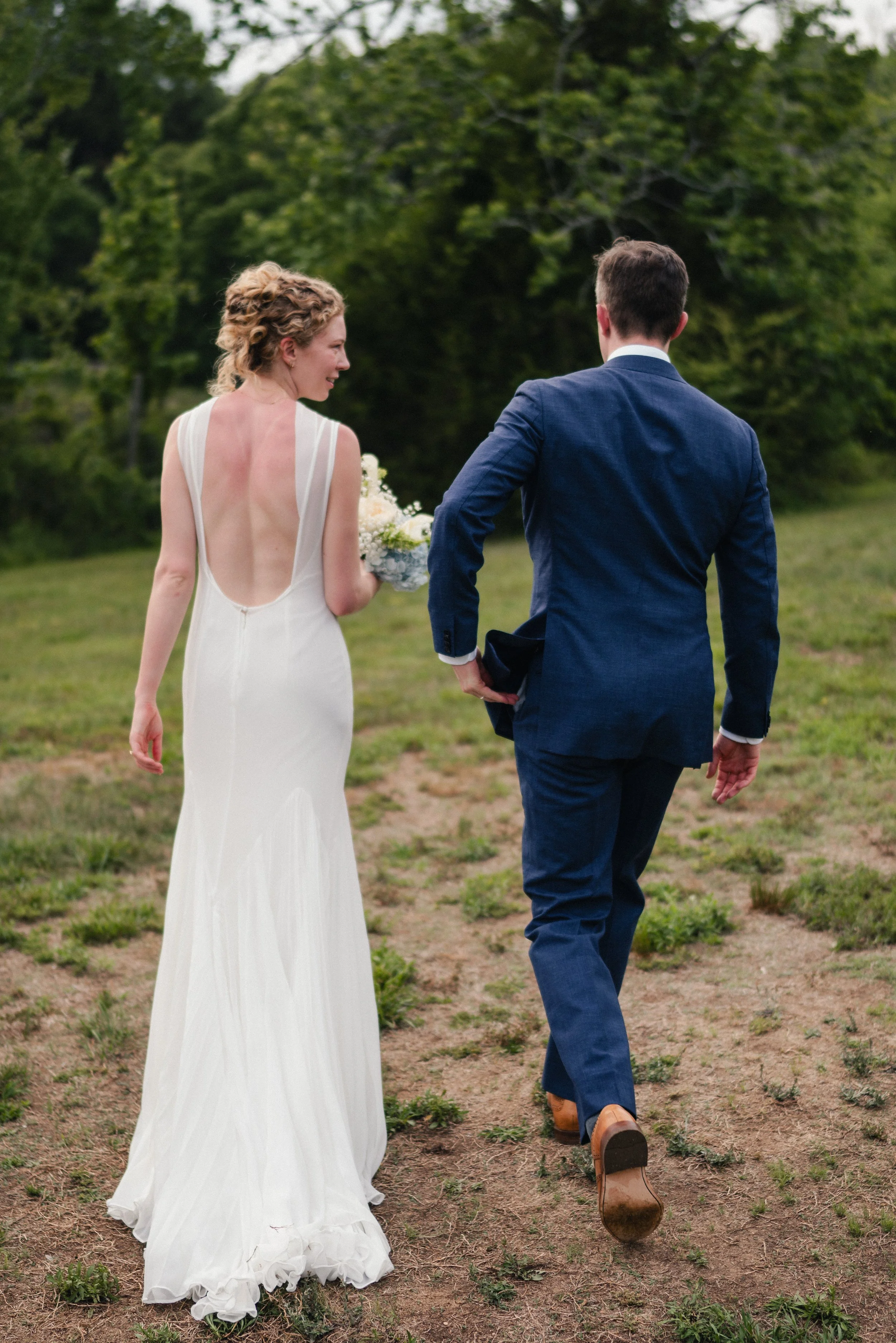 Bride in a white gown walking outdoors holding a bouquet, with groom in a blue suit walking beside her on a grassy area with trees in the background.