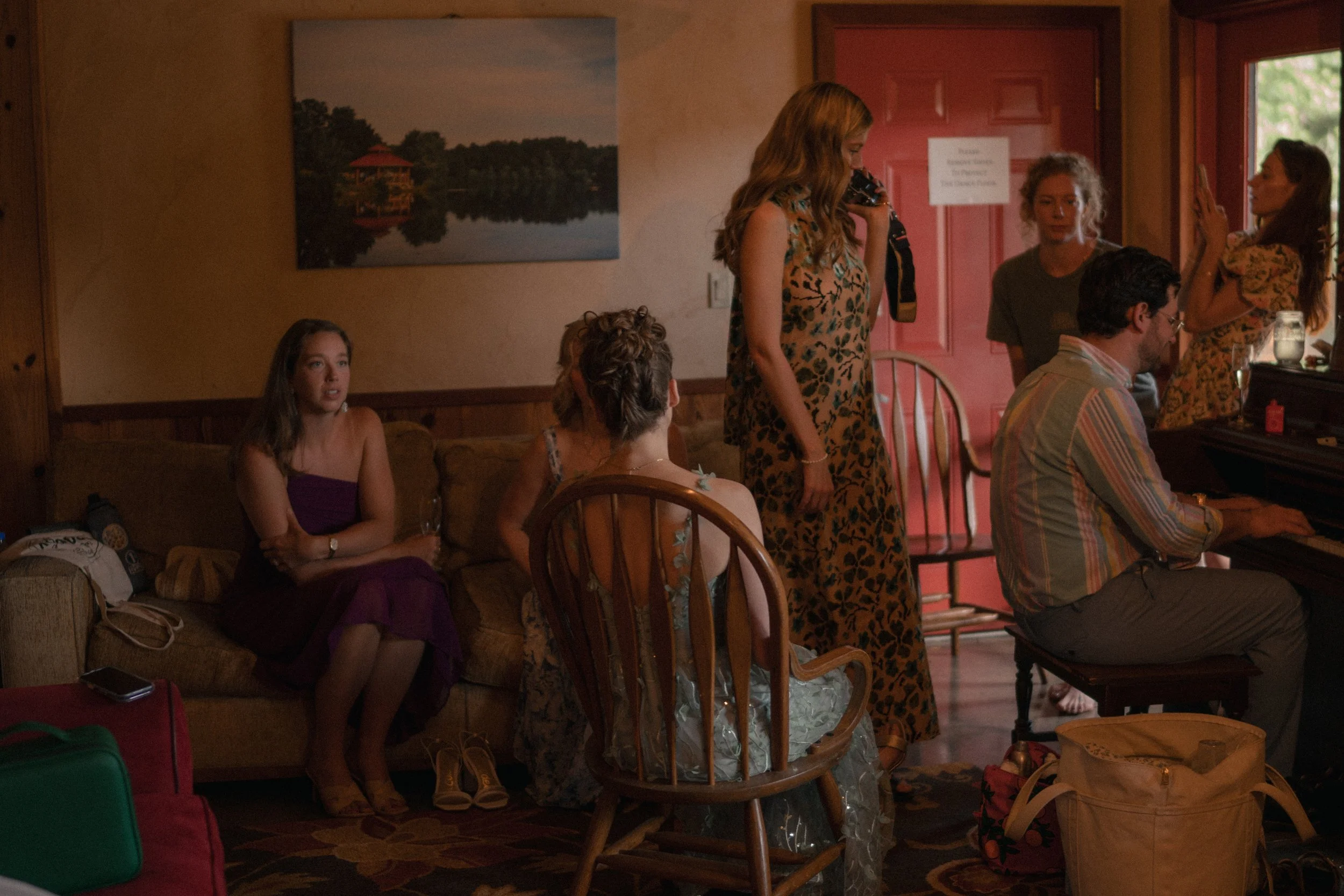A group of young people gathered indoors, some sitting on a couch and others standing, as one girl in a floral dress takes a photo with a camera. A man is playing the piano nearby, and a girl in a purple dress is sitting on the couch looking alert. T