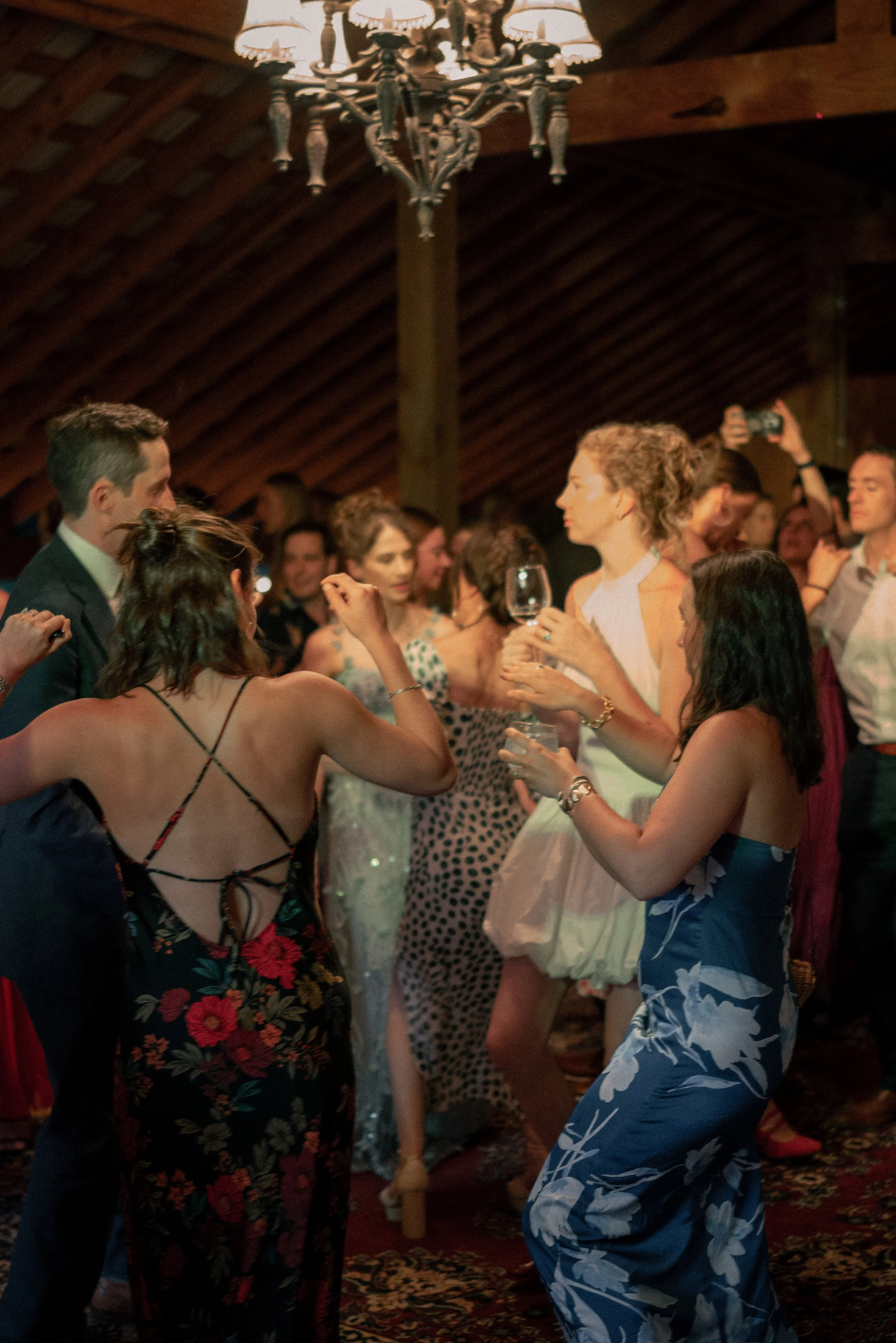 People dancing and socializing at a lively indoor party, with wooden ceiling and a chandelier overhead.