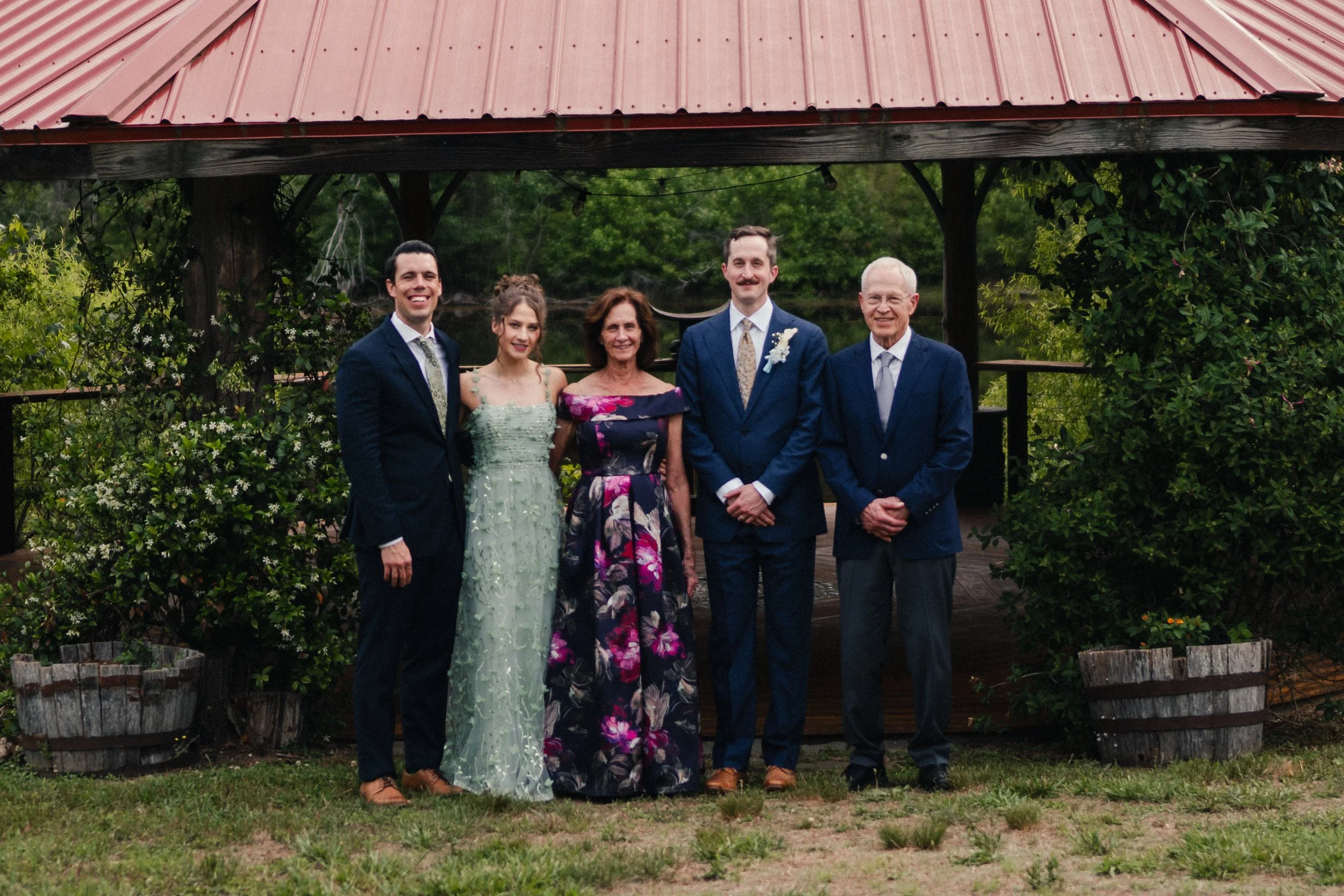 A family of five dressed in formal attire standing outdoors in front of a gazebo with a red metal roof, surrounded by greenery and flowers.